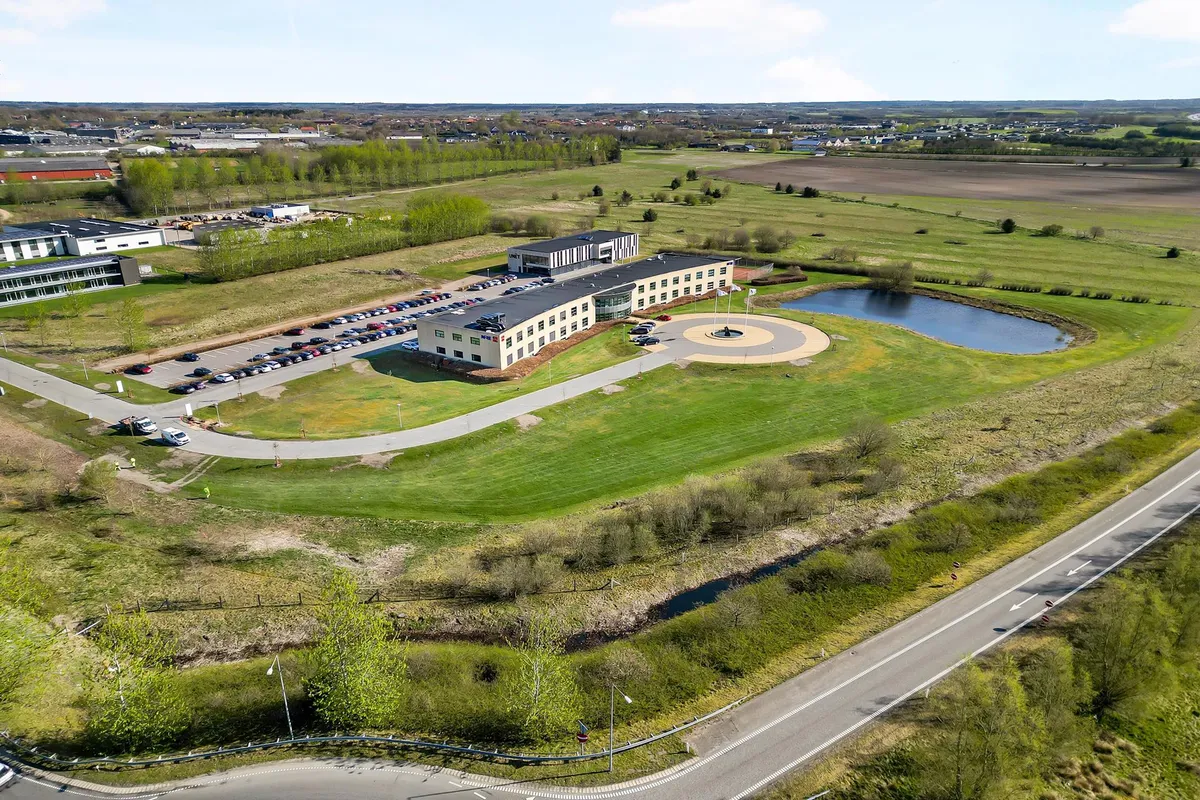 Udsigt over et kontorområde med UNĒ T bygning, parkeringsplads, vand og grønne områder, med vej og en landsby i baggrunden