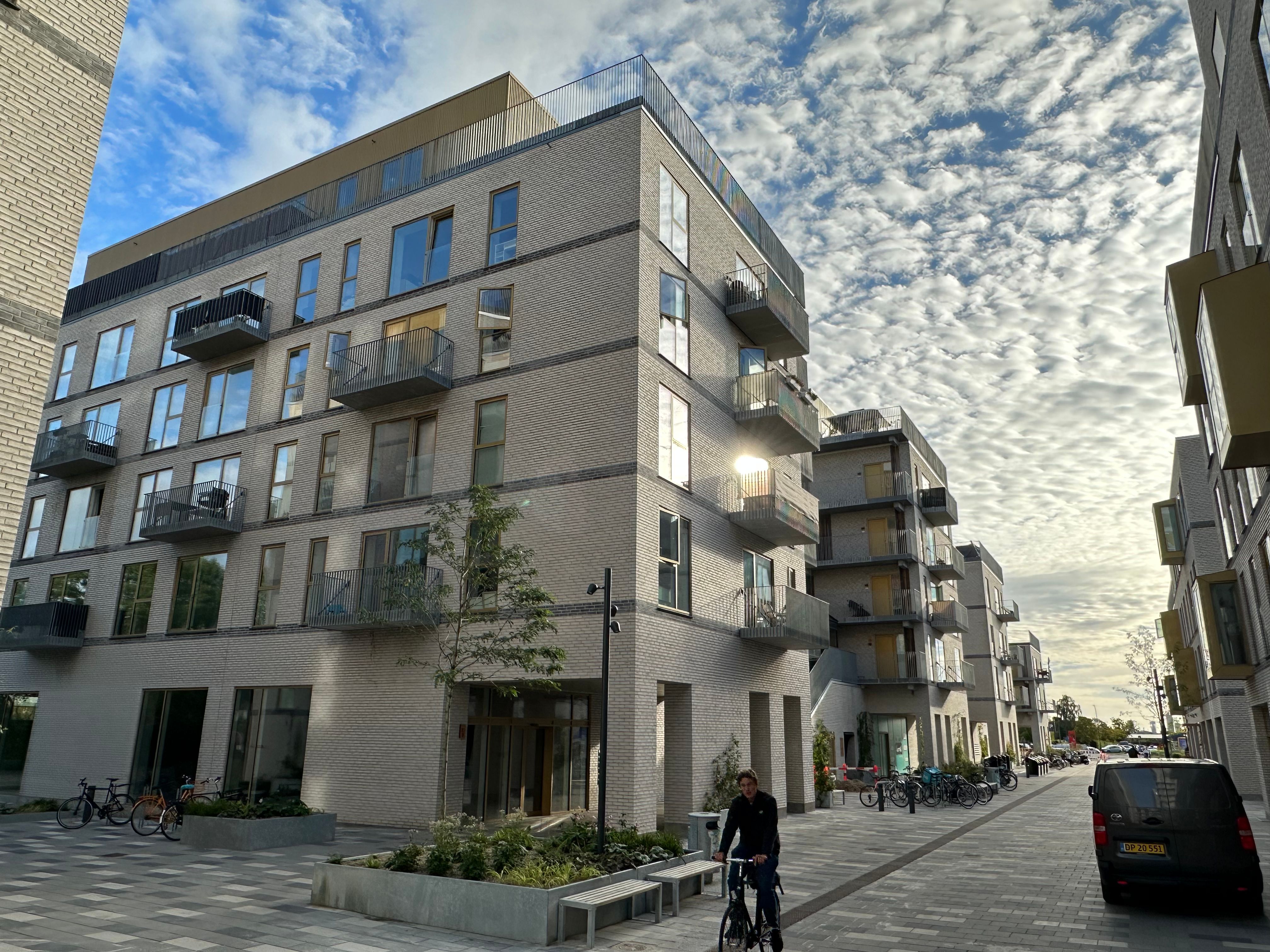 En mand kører på cykel på gågaden foran et højhus med balkoner og udsigt over byen under en delvist skyet himmel