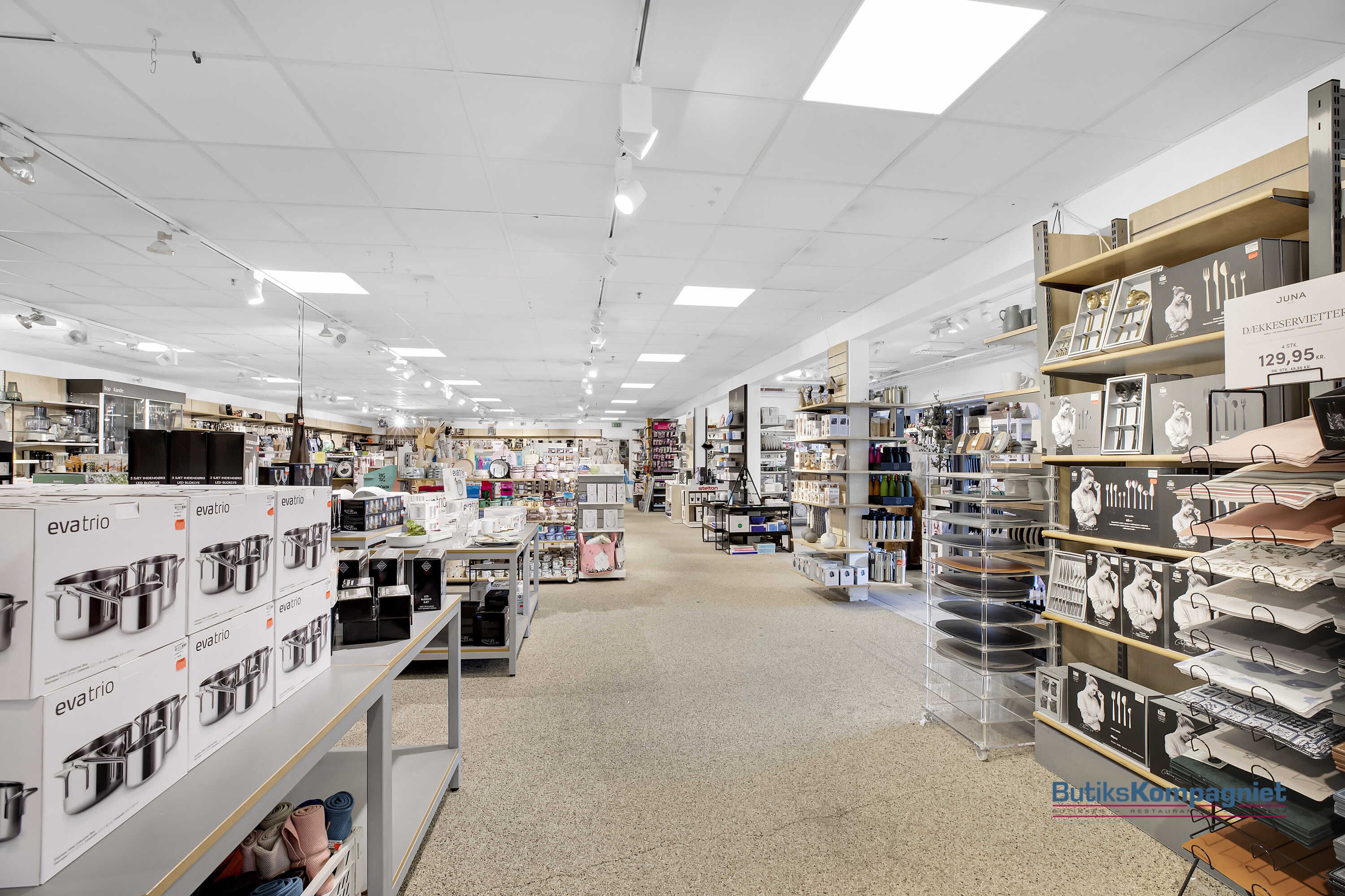 Butik med højt loft og lysegul gulvbelægning, produkter og emballage på hylderne og et mærke på siden med teksten