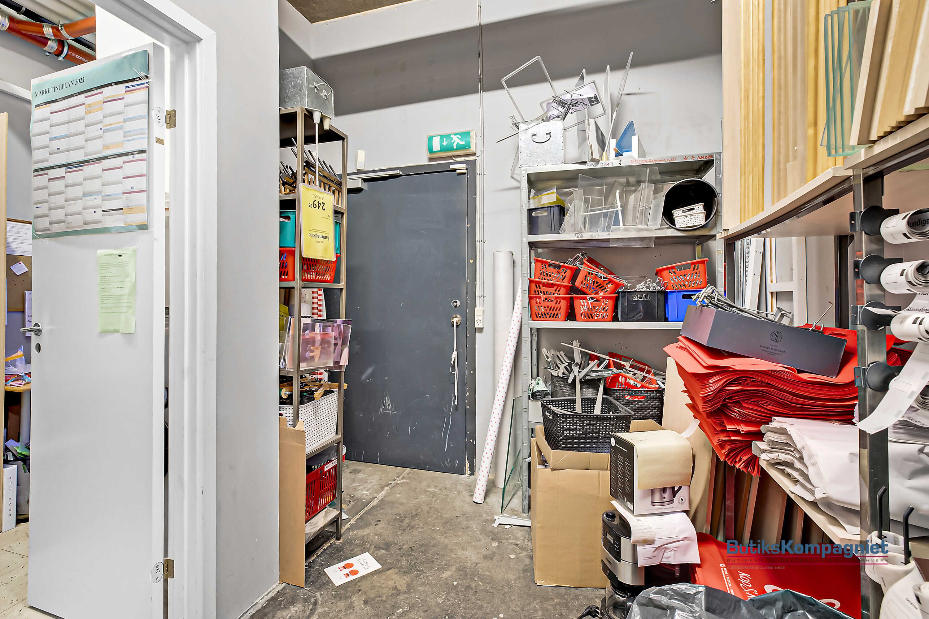 Lager med åbne døre og indvendige hylder, fyldte med redskaber og emballage. Et rødt kurvstativ er placeret på en hylde.