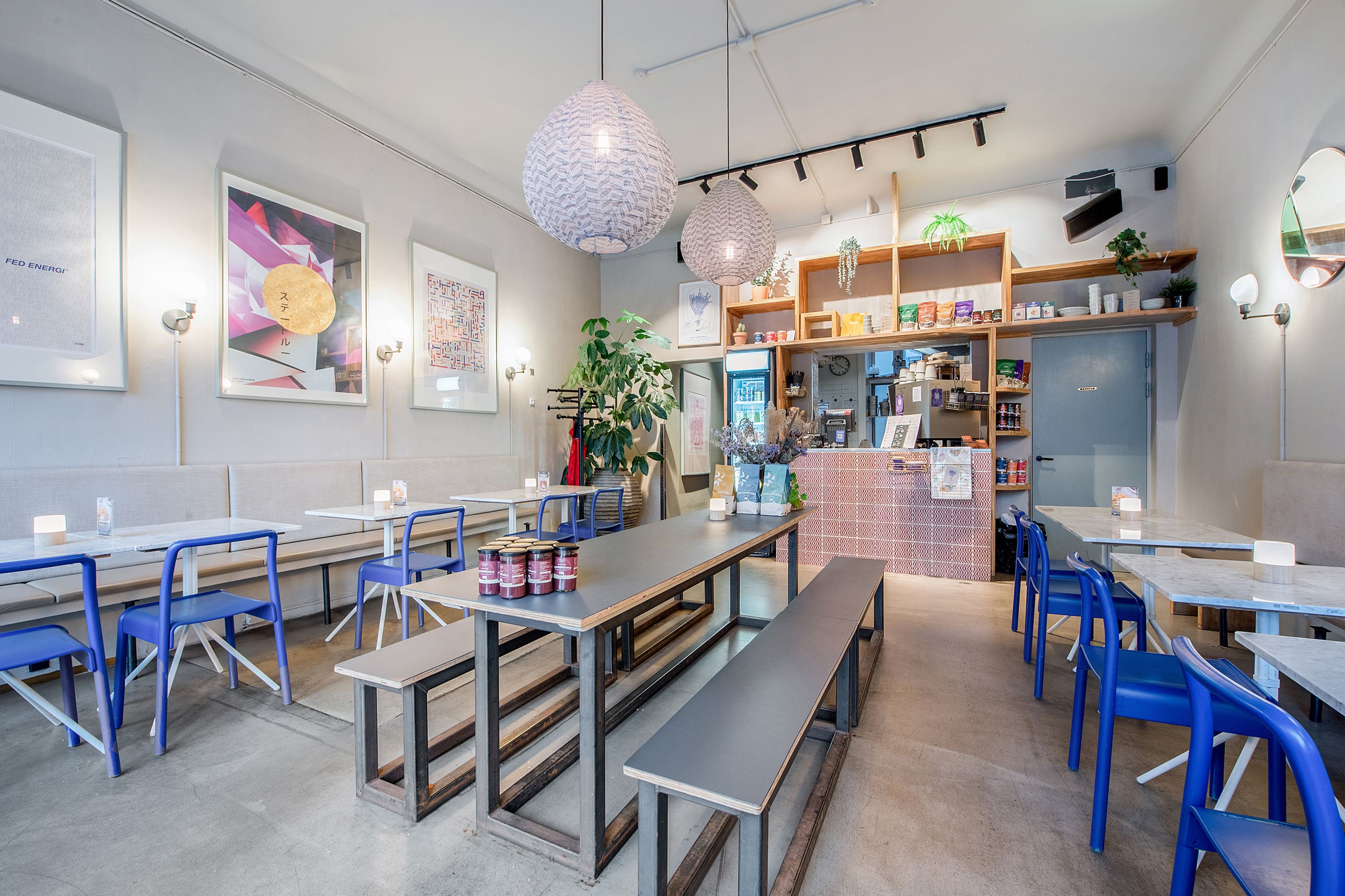 Moderne café med højt loft, pæne bordopstrækninger og lyse loftslampe. Indeholder store billeder og store planter.