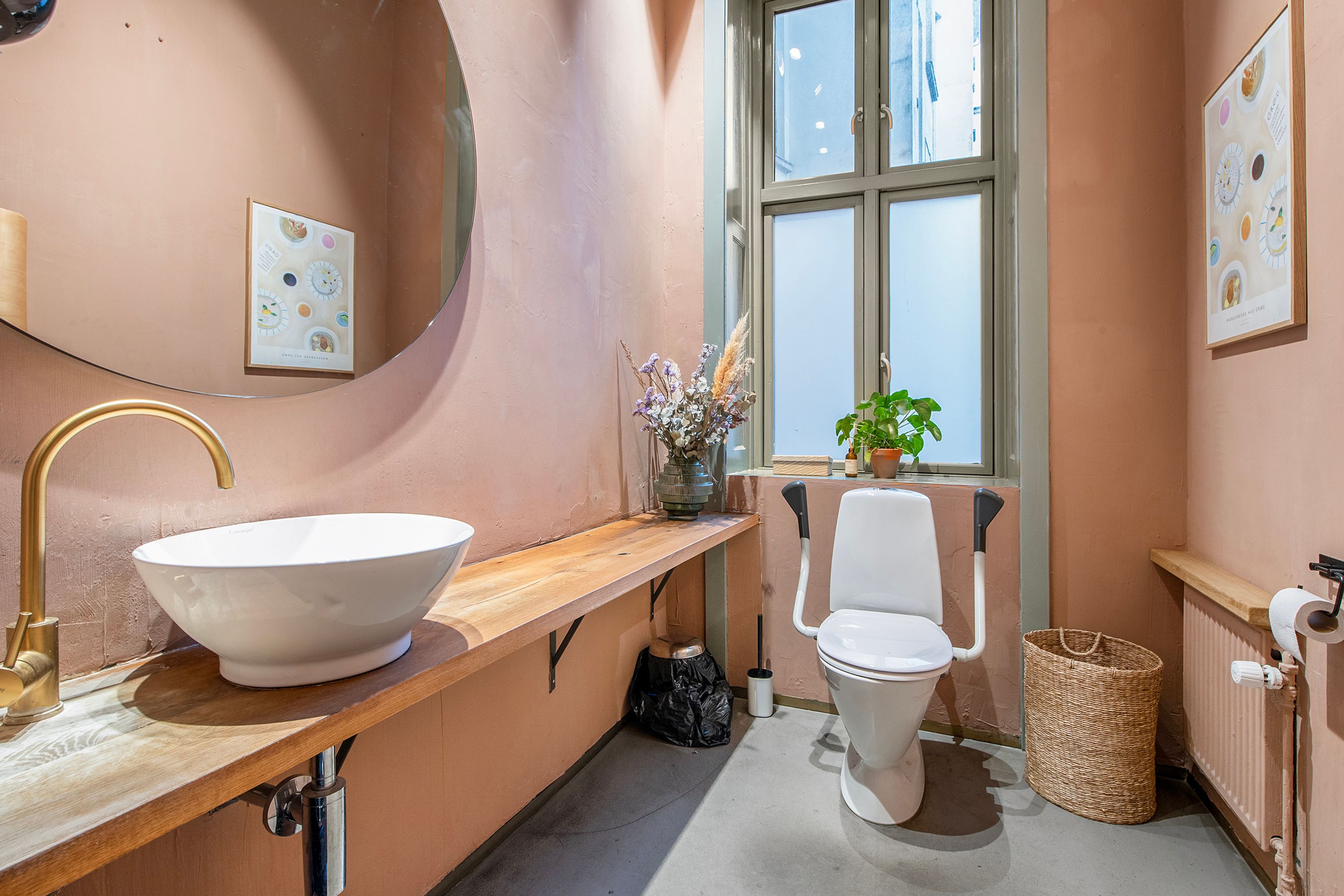 På en hyggeligt indrettet badeværelse ses et hvidt, rundt tøjvask, toilet med håndtag, plante i vase og en lyse pink farvede