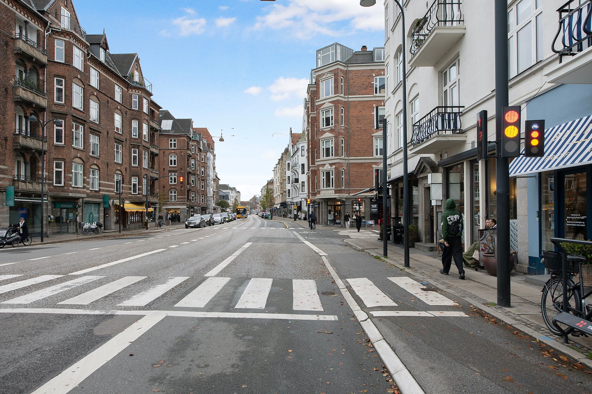 En bred gade i Hellerup med fortov, zebraovergang, biler og folk, der går og cykler i efterårssol og blå himmel