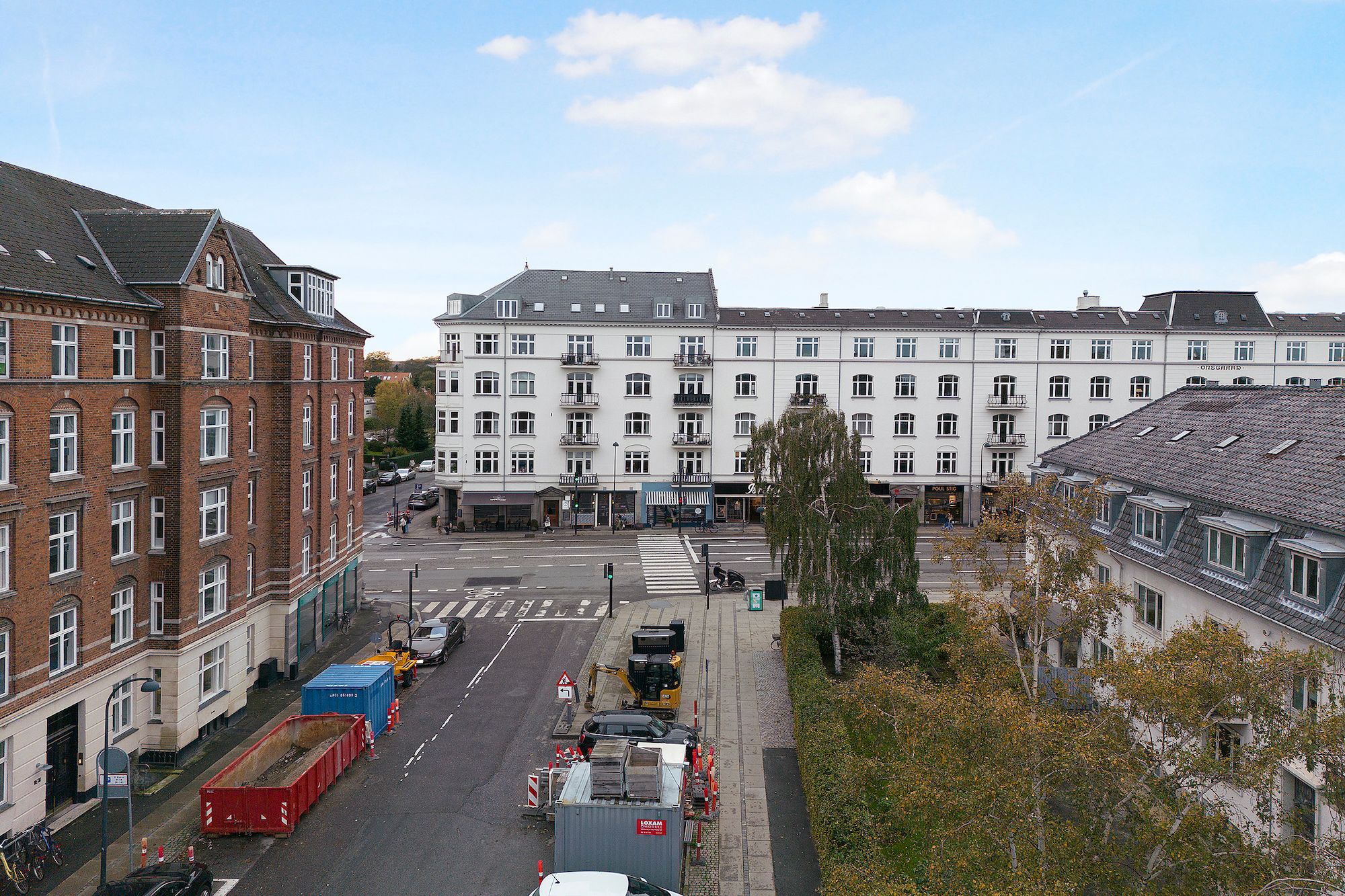 Vejkryds med lejemål, containere, byggemaskiner og grønne træer og buske.