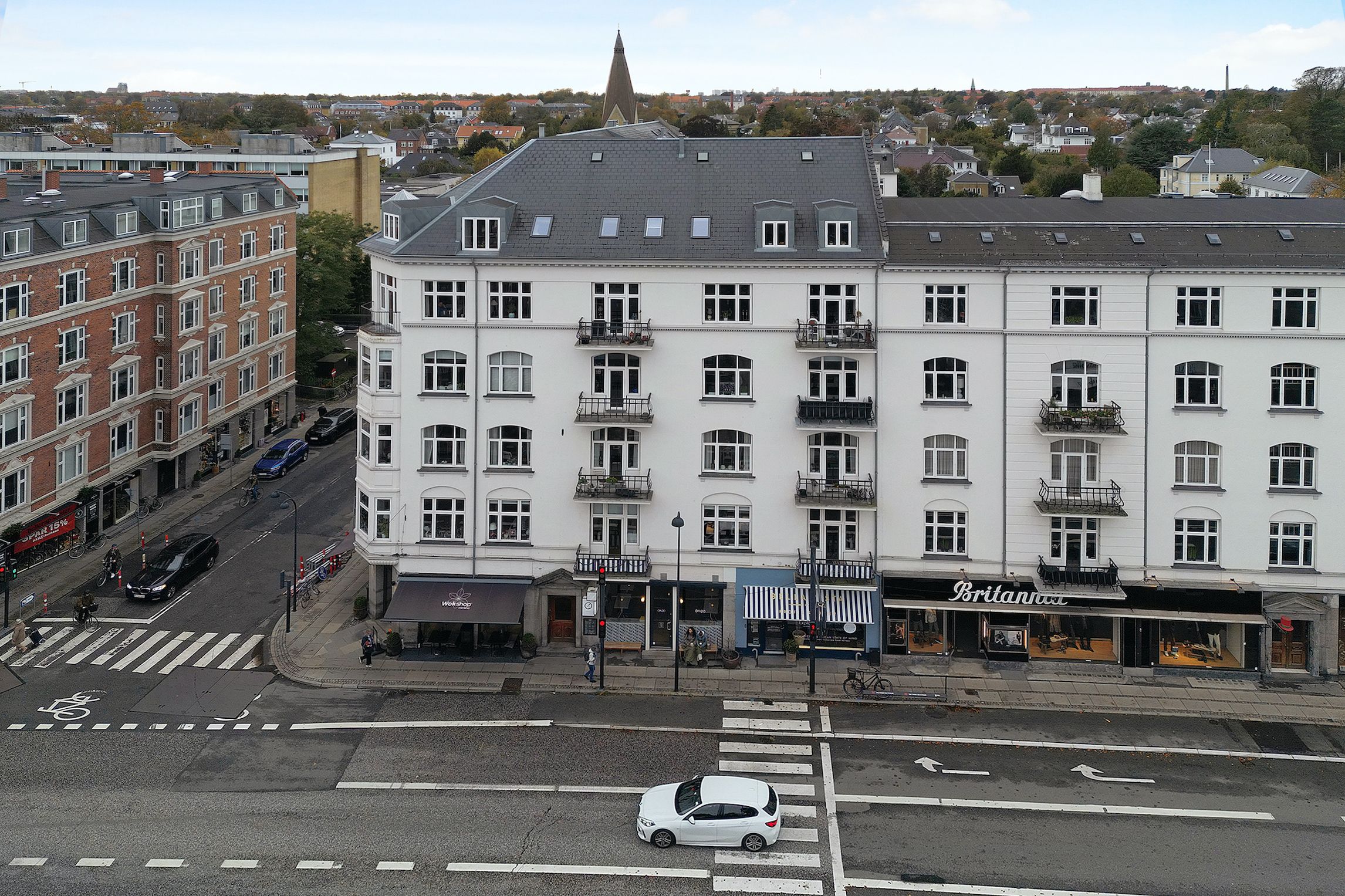 Vejkryds med en hvid hatchback bil og krydsende fodgængere, med strandvejen i baggrunden og butikkerne Brittannus og Vin og