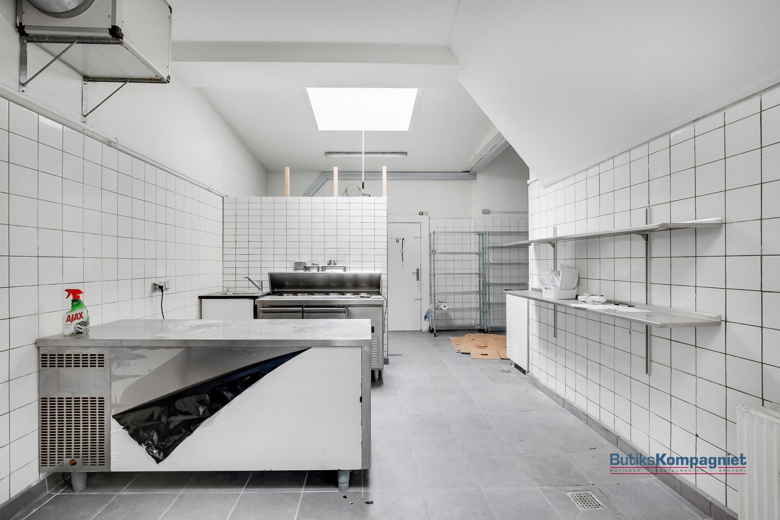 Et funktionelt køkken med højt til loftet, lysegulv, flere apparater og skrald. Væggene er hvide og flisede, og der er et