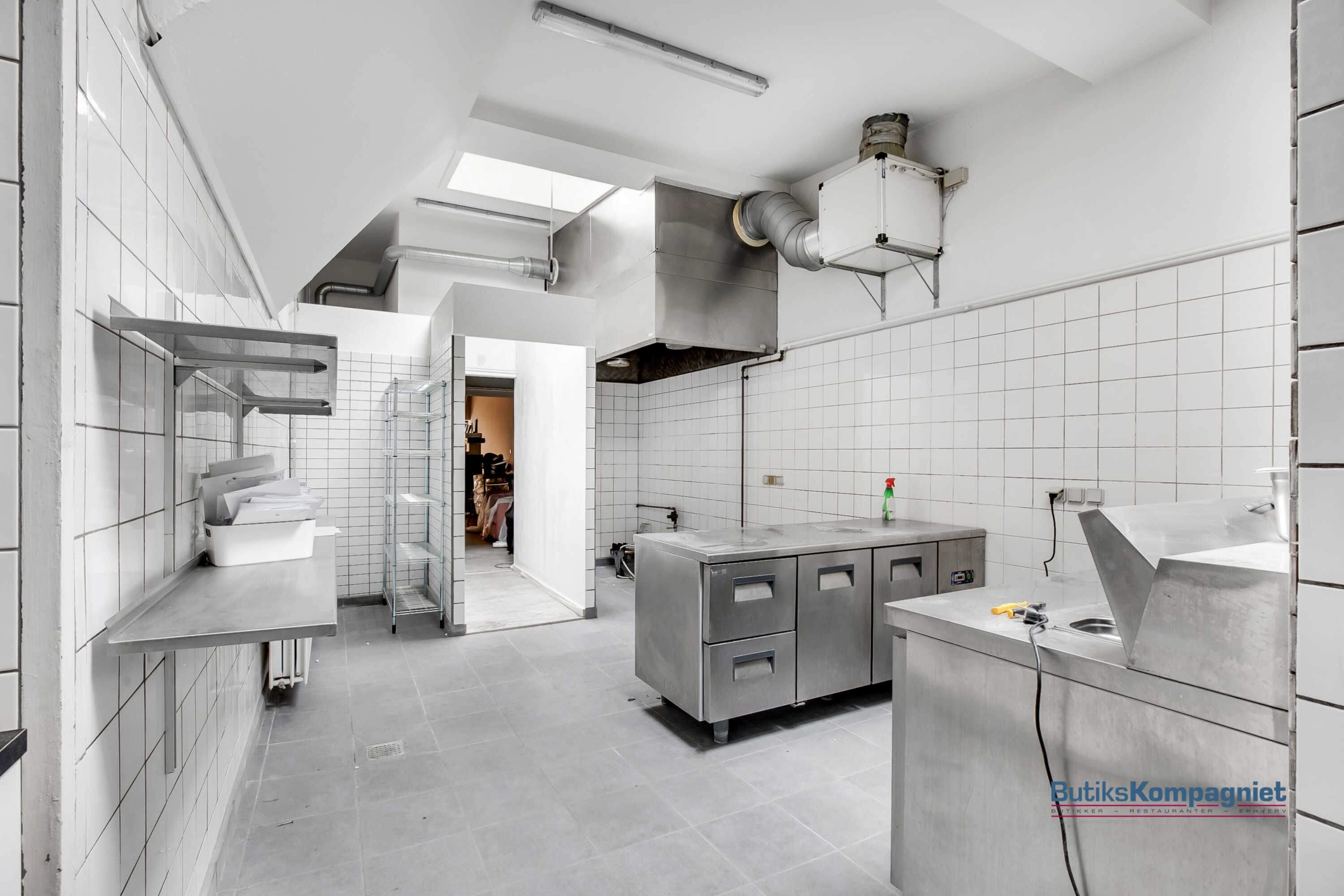 Et rum med hvide vægge og gulv, der er udstyret med en række metalhylder og en stor metalbord med to vaskebassiner og en