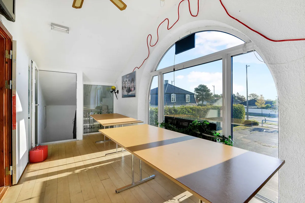 Indvendigt kontor med to lange borde og lyse gulv. Væggene er hvide, og der er en stor bueformet vinduesglas. Et billede er