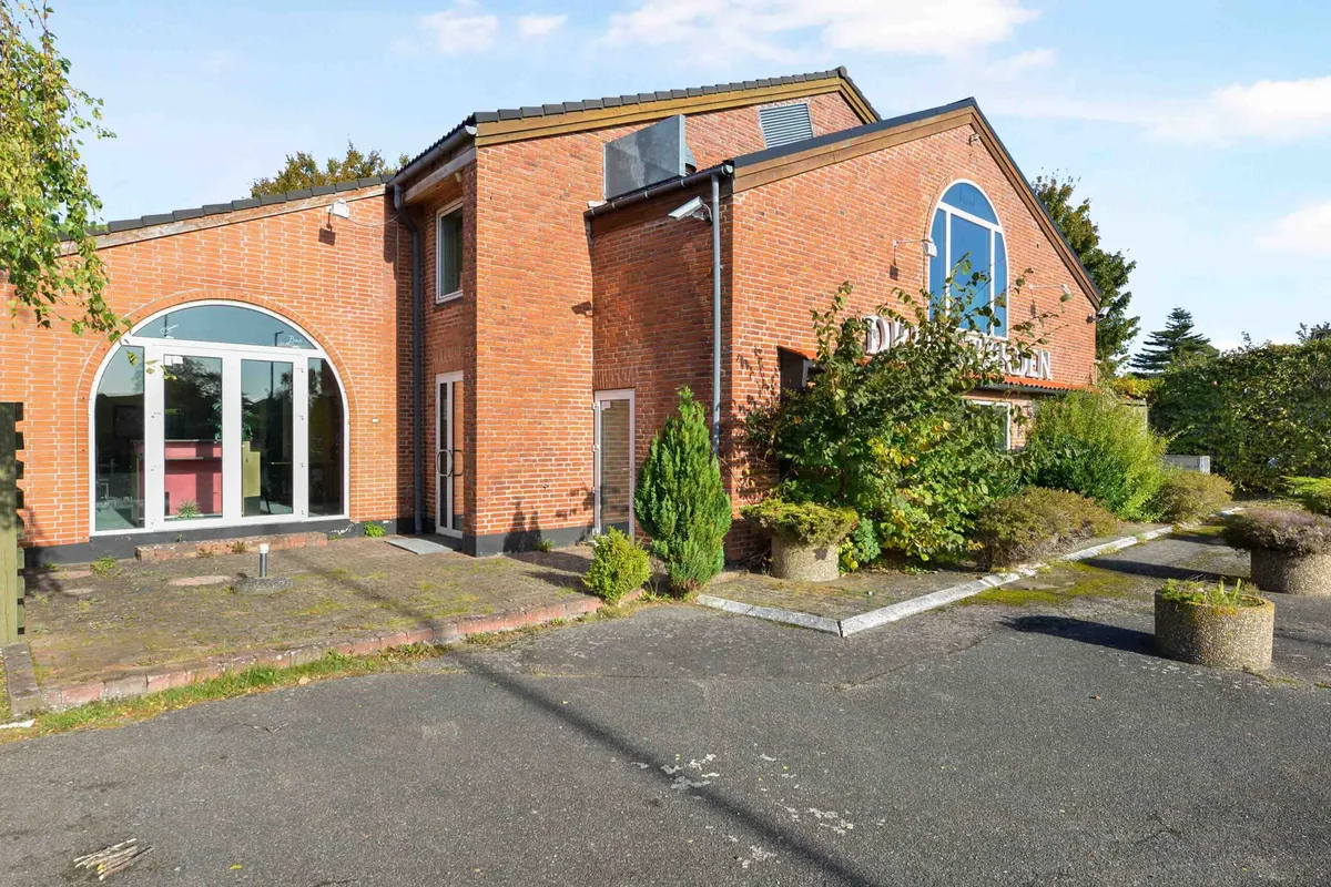Rødt murstenshus med to vinduer og en stor glasdør, foran er to cementplanter og en brolægget indkørsel, der fører til en