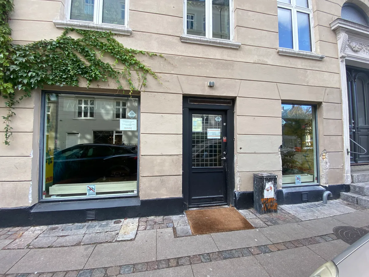 Erhvervslokaler med glasruder og dør, hvor en bil er reflekteret i vinduet. En væg er dækket af grønne planter og en