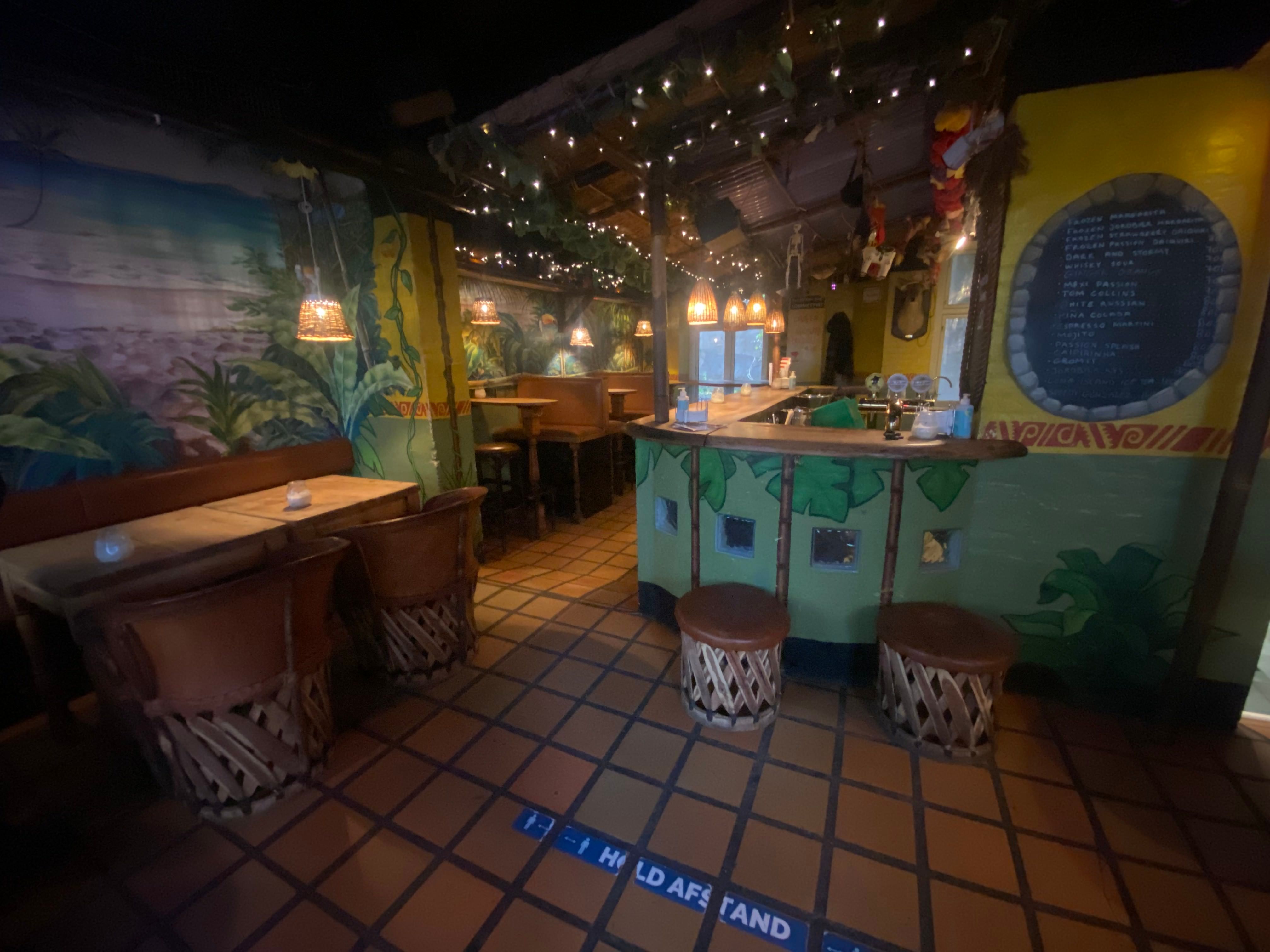 En bar med stolpe, stolpe, borde og stolene og en udsmykning med planter og lysekroner og menuen på væggen