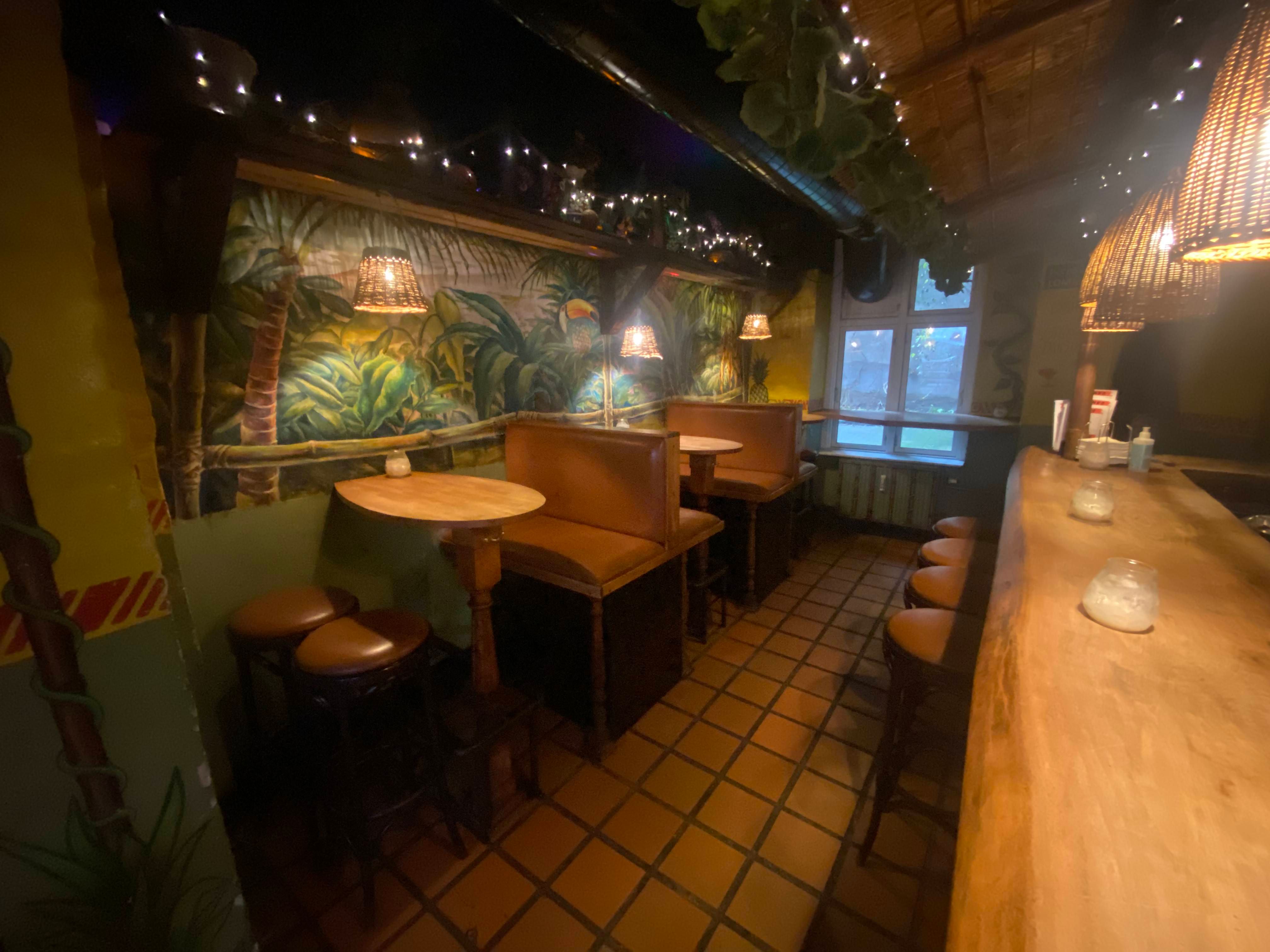 Indretning med brunlæderstole og runde borde, lysekroner hængende fra loftet, vægdekoration med tropiske motiver og træbord