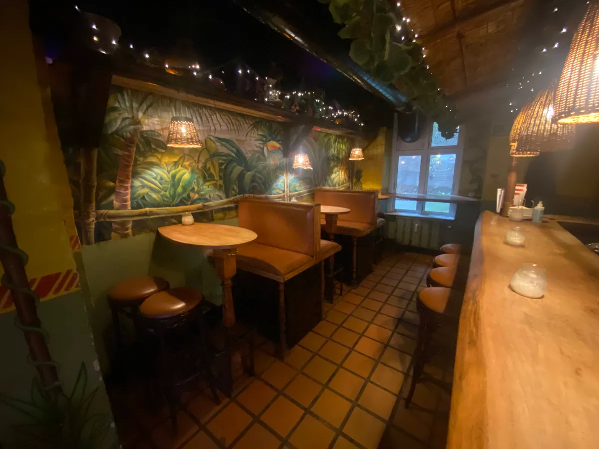 Indretning med brunlæderstole og runde borde, lysekroner hængende fra loftet, vægdekoration med tropiske motiver og træbord