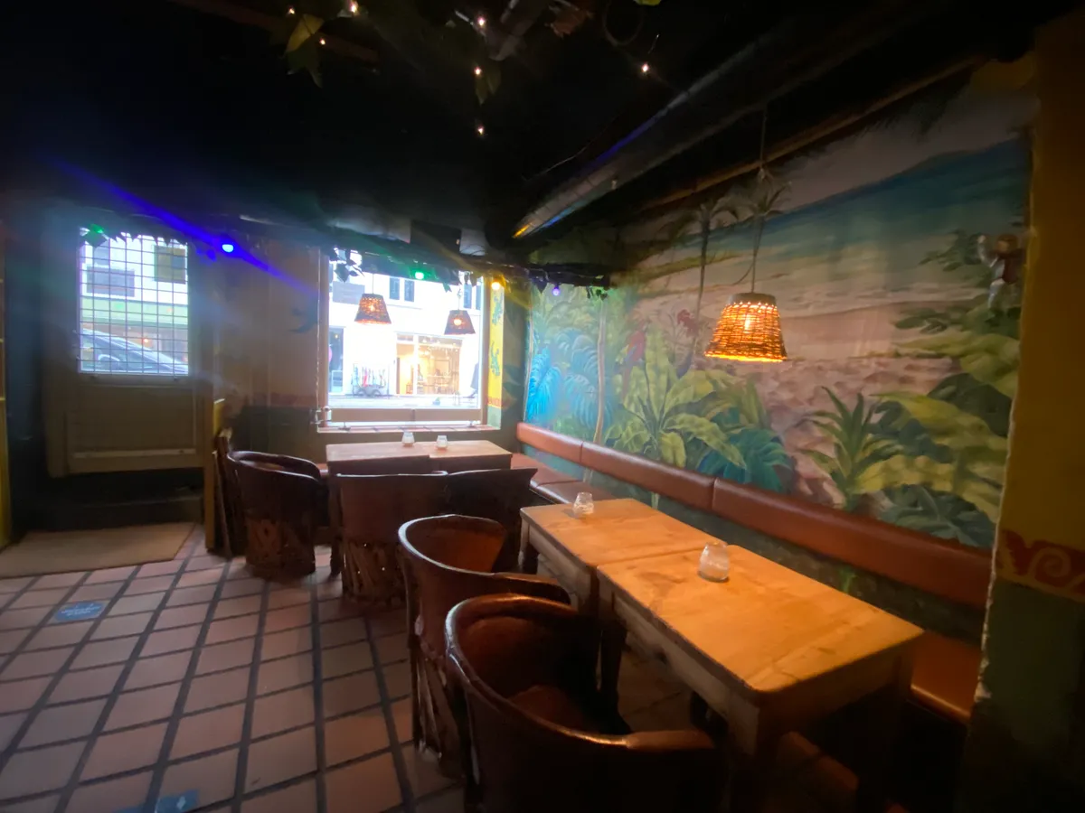 Lystfuld restaurant med lyse kroge og lange træborde samt malerier på væggen med tropiske motiver