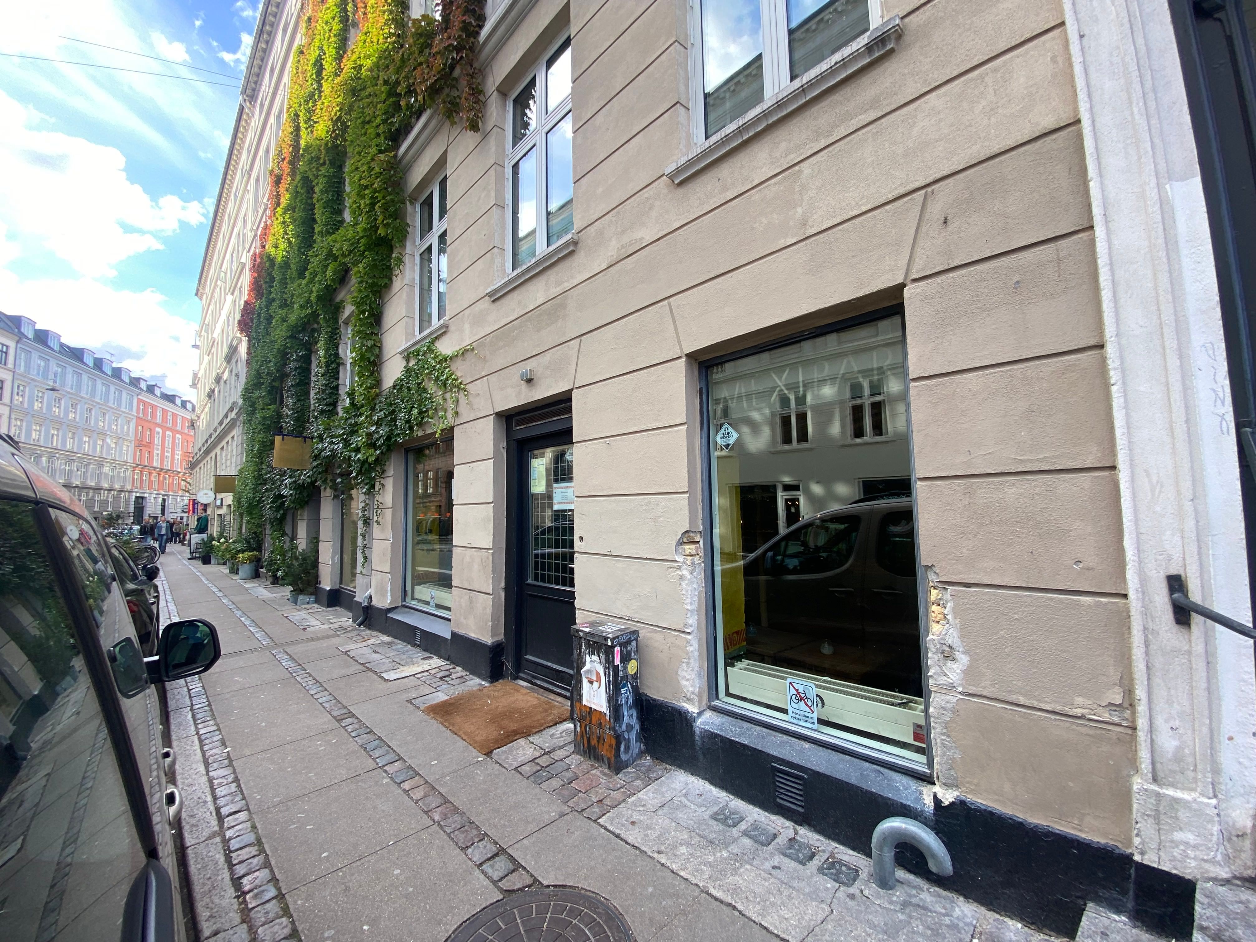 En stor bygning med grønne planter og en høj skorsten står på en gade med en bilparkeret bil og en tyk skorsten