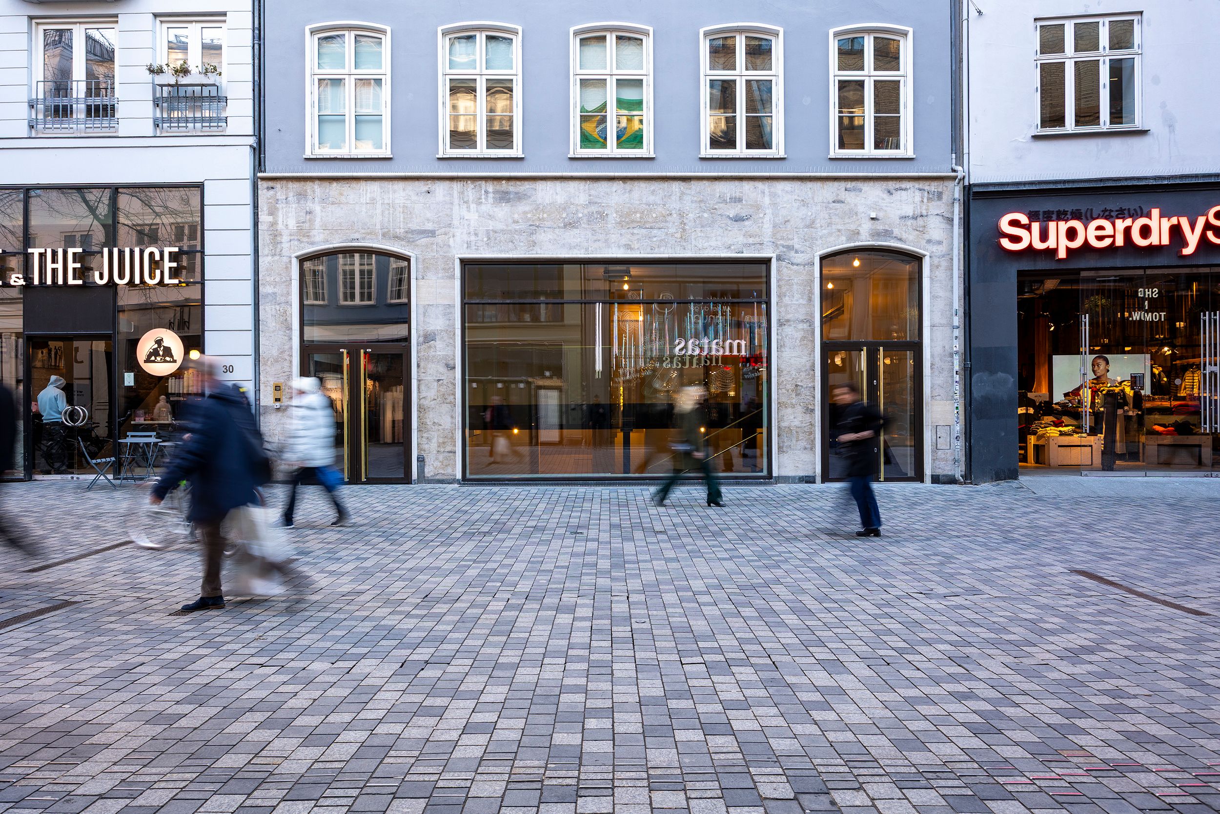 Mennesker går forbi Superdry og The Juice, der er butikker i baggrunden, og der er flere mennesker i midten. Butikkerne