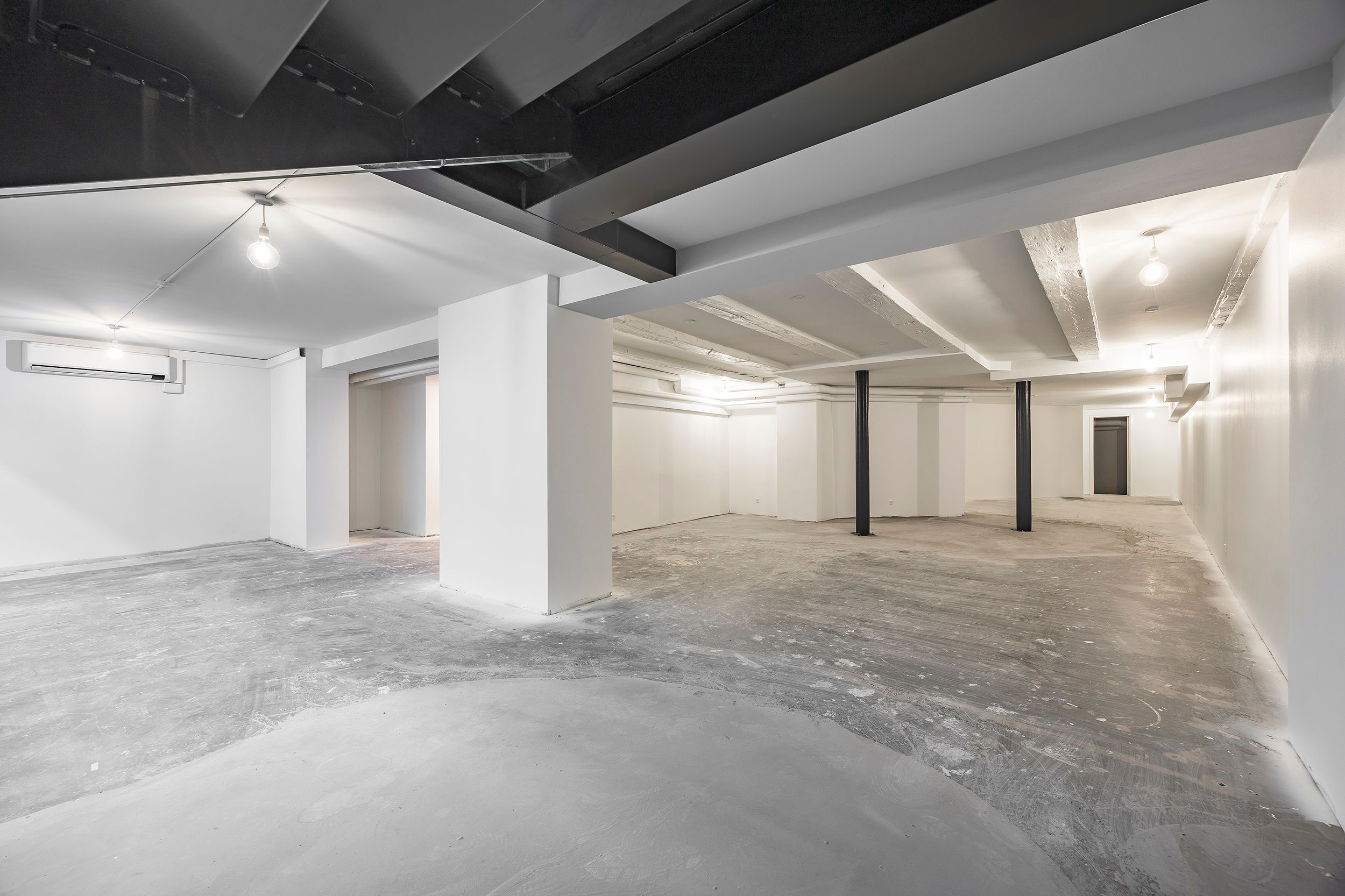 Store tomt rum med cementgulve, flere hvide vægge, og lysekroner. Loftet er højt og er i sort. Der er et klimaanlæg monteret