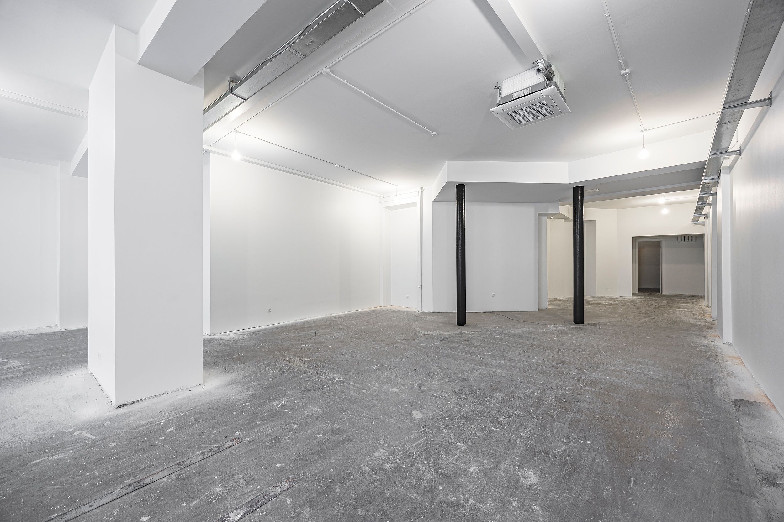 To store hvide søjler og to mindre højere søjler står i et tomt lokale. Væggene er hvide, og gulvet er gråt og ujævnt.