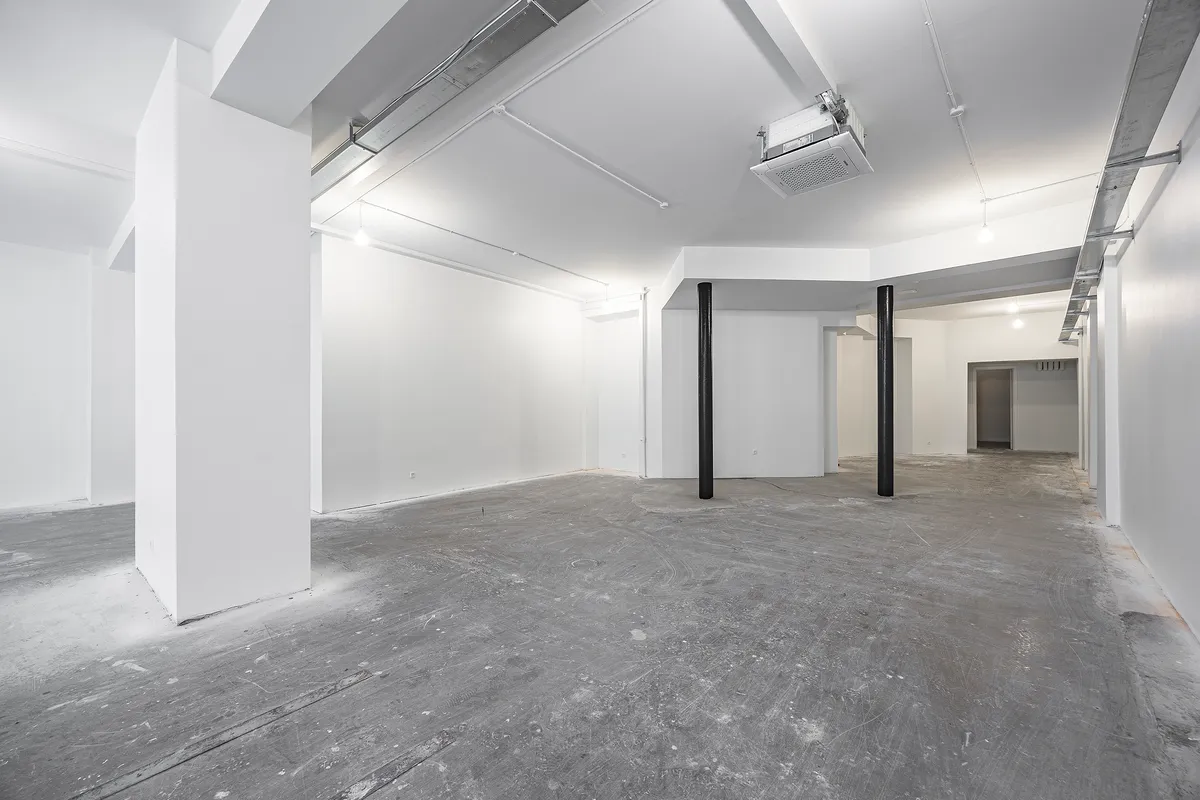 To store hvide søjler og to mindre højere søjler står i et tomt lokale. Væggene er hvide, og gulvet er gråt og ujævnt.