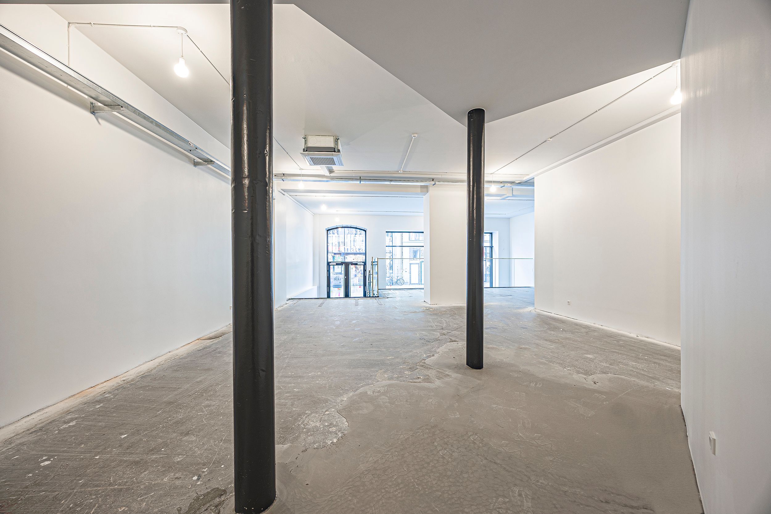 Foto af et tomt rum med hvide vægge, højloft og to stålsøjler i midten og to døre i baggrunden
