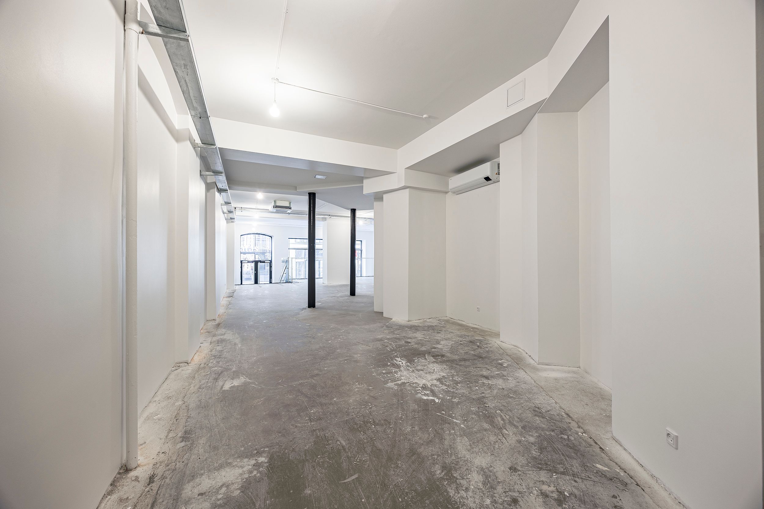 En tom lejlighedsgang med hvide vægge, gulve og loft. Der er en metalrør i loftet og et klimaanlæg i væggen. Gangen har et
