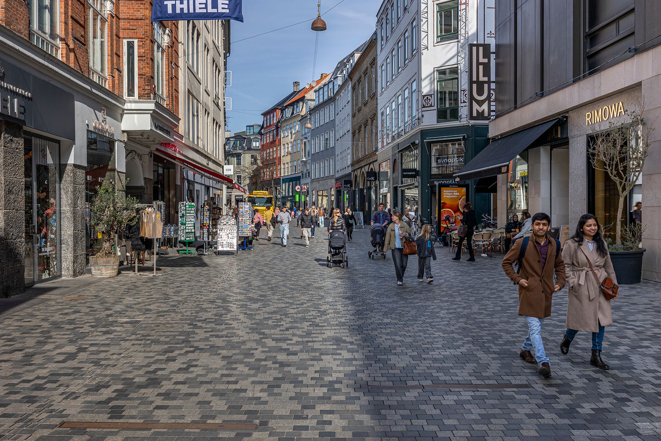 En travl gågade med folk, butikker, en børnevagt og en bus, samt en kvinde og mand, der går mod hinanden