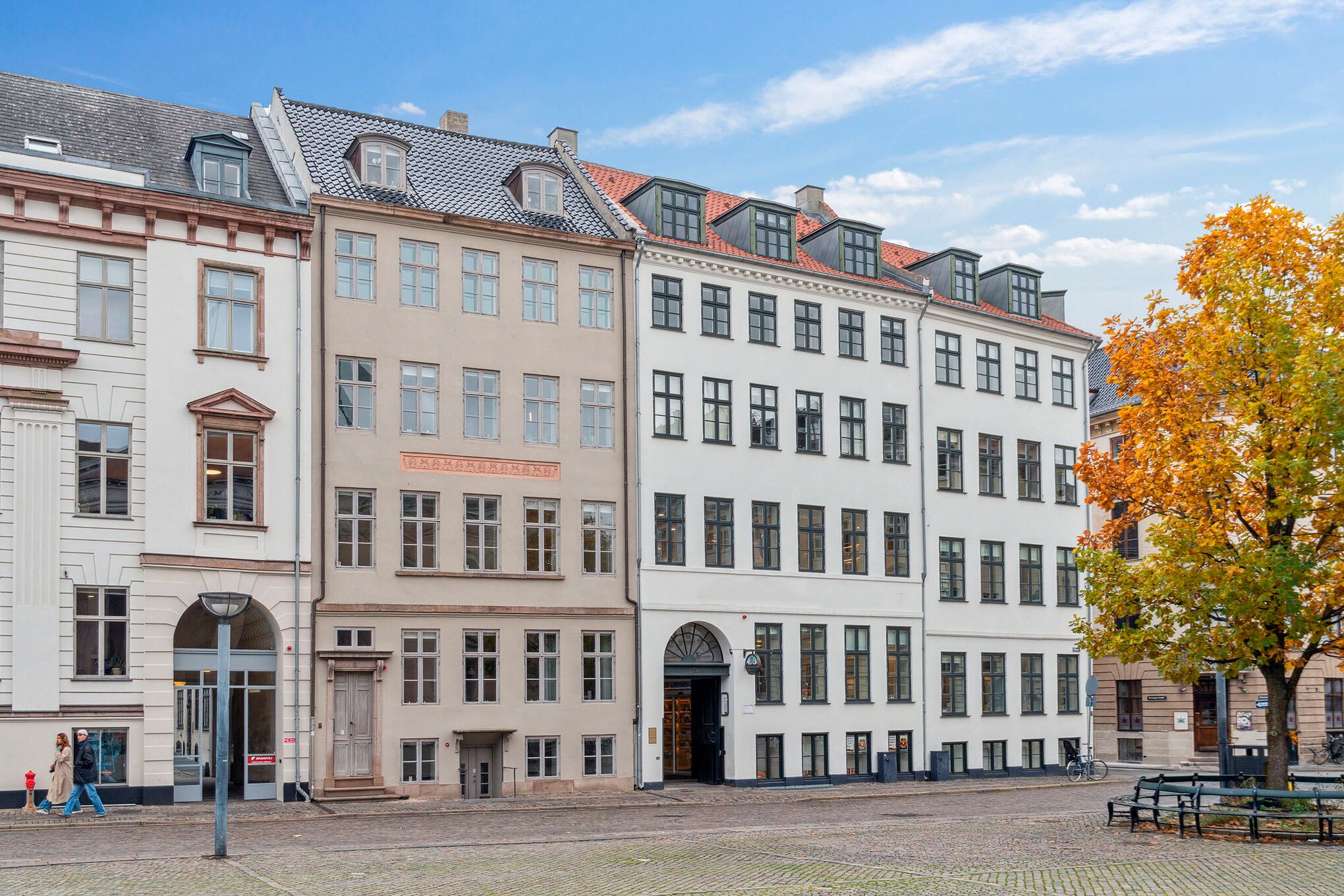 Man går forbi tre bygninger med højde og slanke vinduer og en lille skovetræ i forgrunden