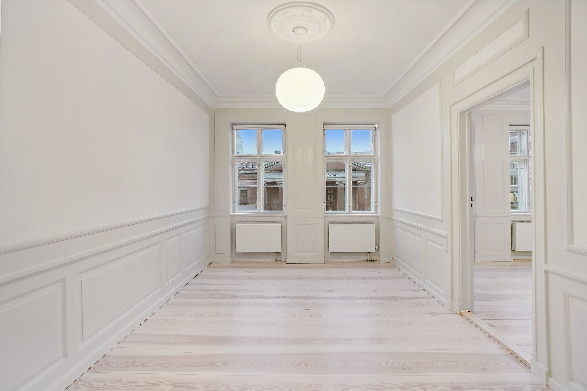 En stor og lyse hall med lysegult trægulv, to store vinduer og et rundt loftslys