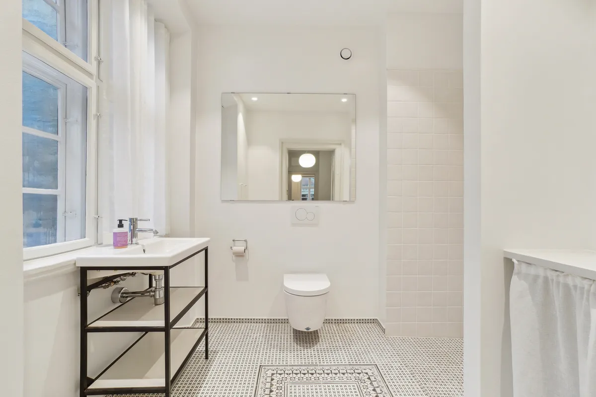 Moderne badeværelse med hvidt toilet, en stor spejl, hvidt skab, hvidt badekar og en vinduesside med hvidt gardin. På