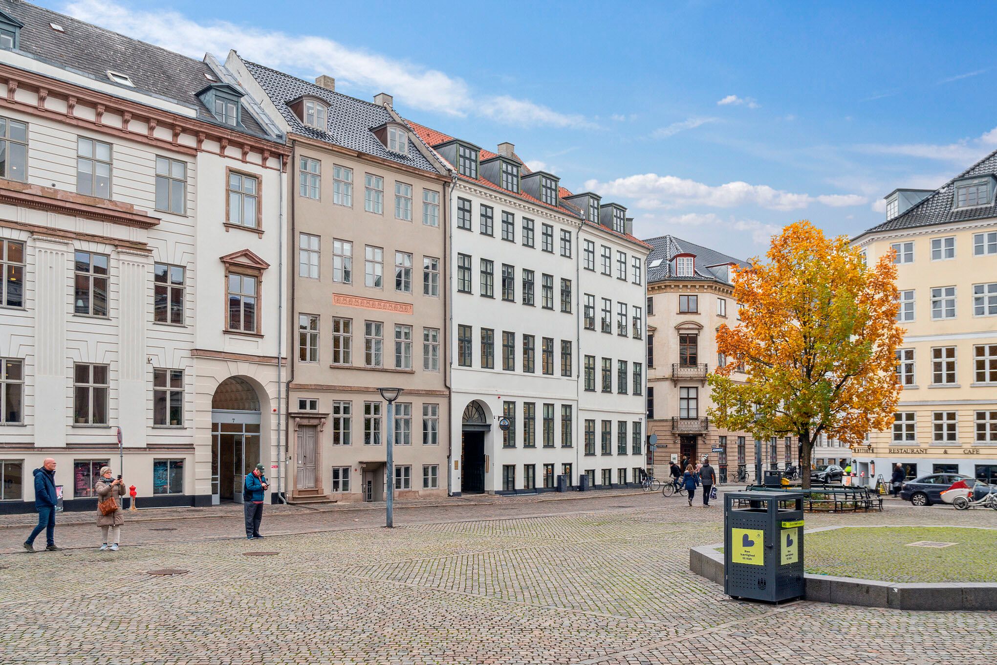Mennesker går på Nytorv med udsigt over Københavns Byret og en stor træ i centrum. Man ser en skulptur og en skraldespand