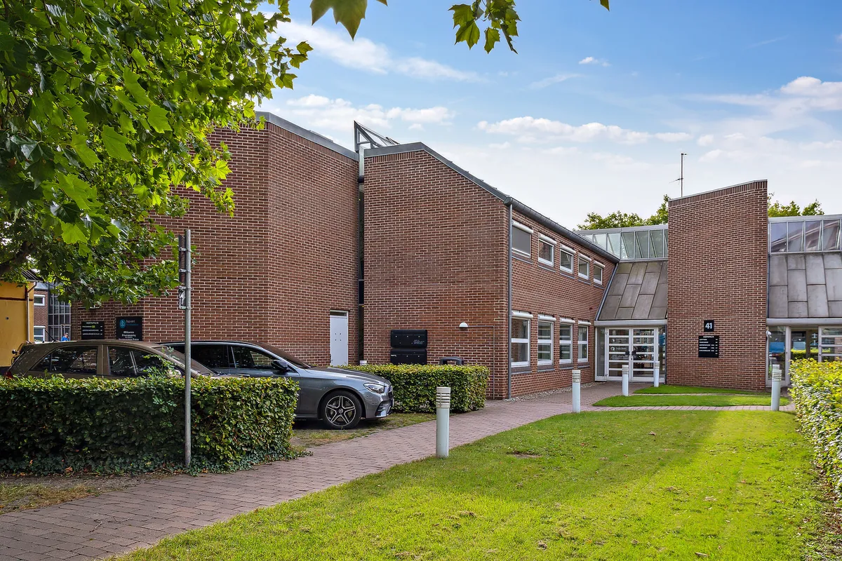 To biler er parkeret på en asfaltvej udenfor et rødmurstensbygning med glasruder og nummer 41. Buske og græsser er plantet