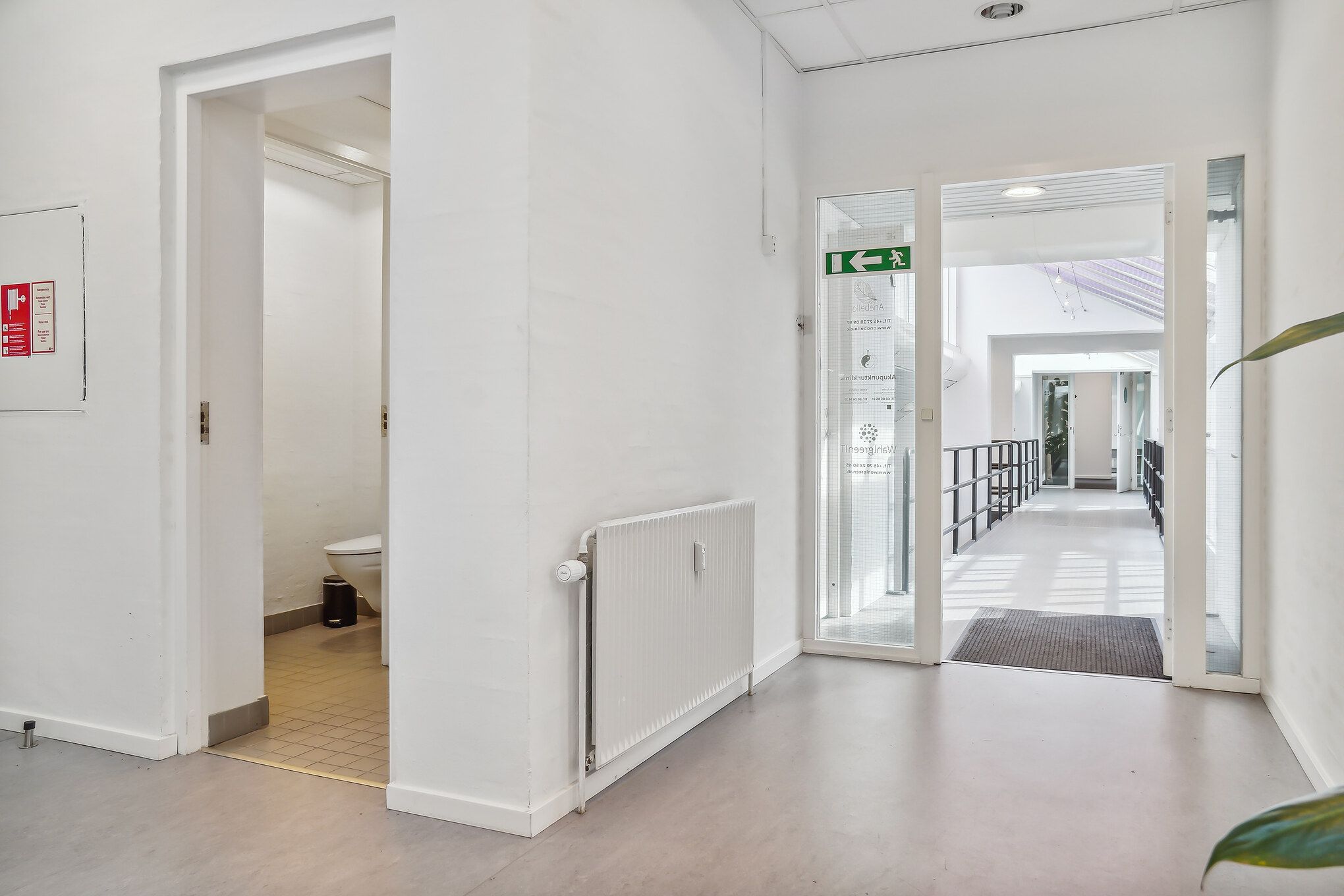 En møbleret toiletrum med toilet, affaldskurv og radiator samt adgang til en korridor med glasdøre og et loft med indbygget