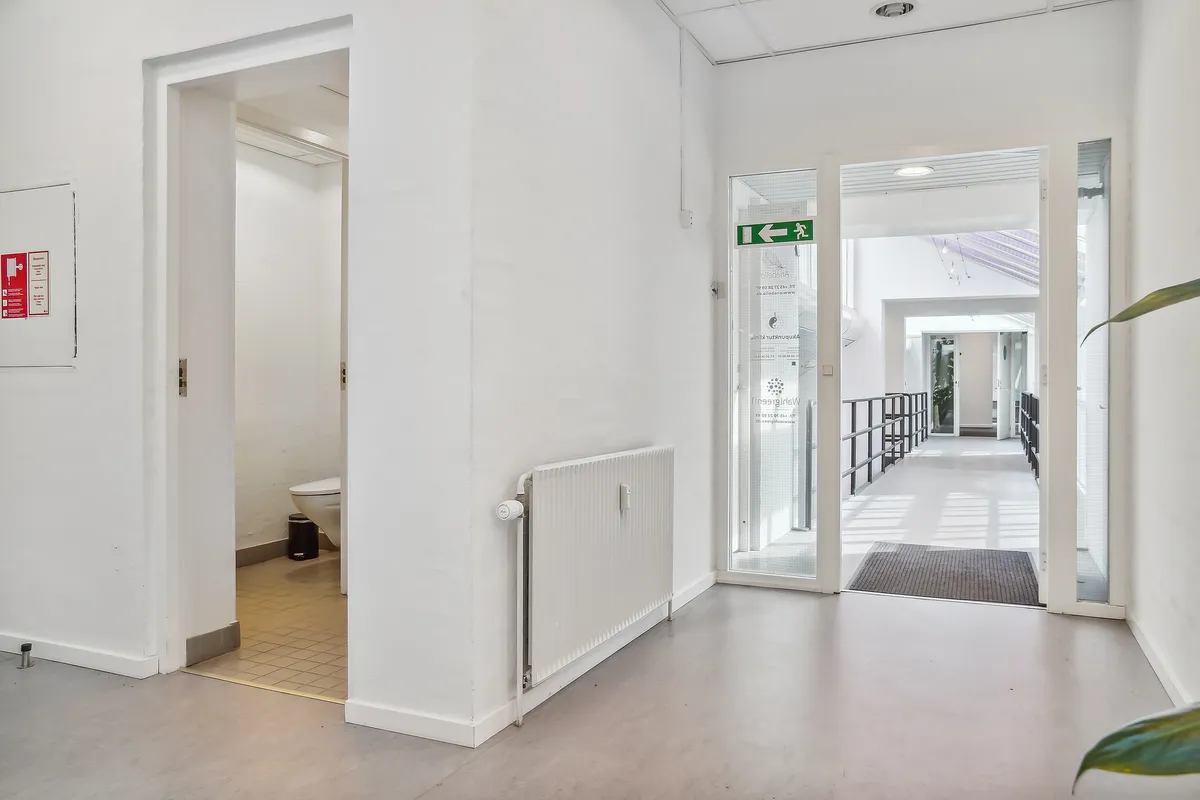 En møbleret toiletrum med toilet, affaldskurv og radiator samt adgang til en korridor med glasdøre og et loft med indbygget