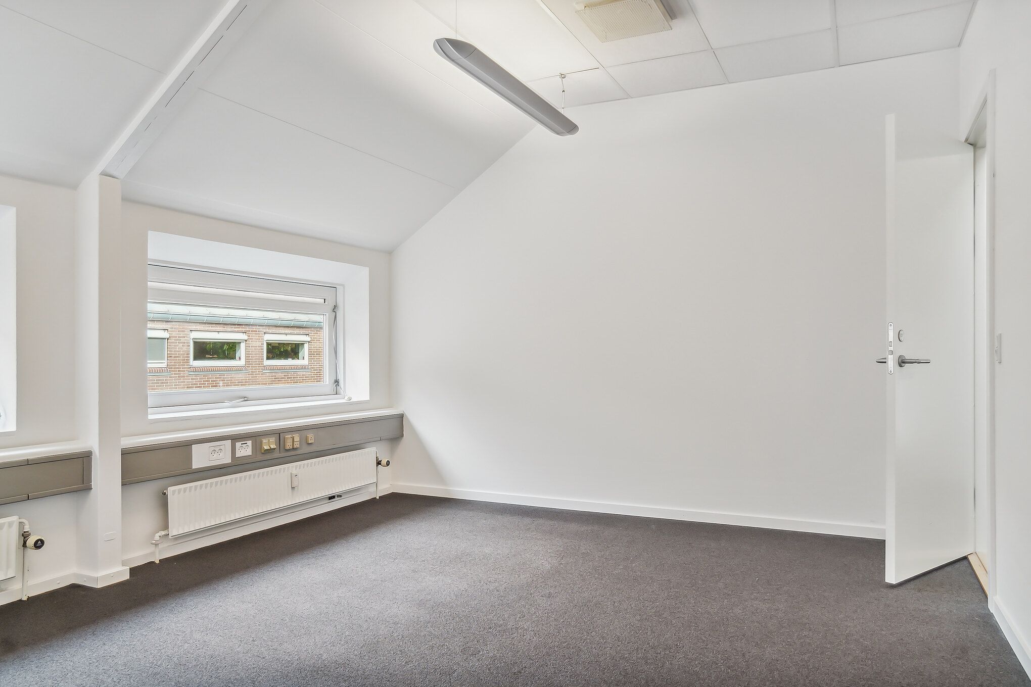 Lyserum med gulvtæppe og radiatorer langs væggen. Vindue med udsigt til bygning med røde mursten og to mindre ruder. Åben