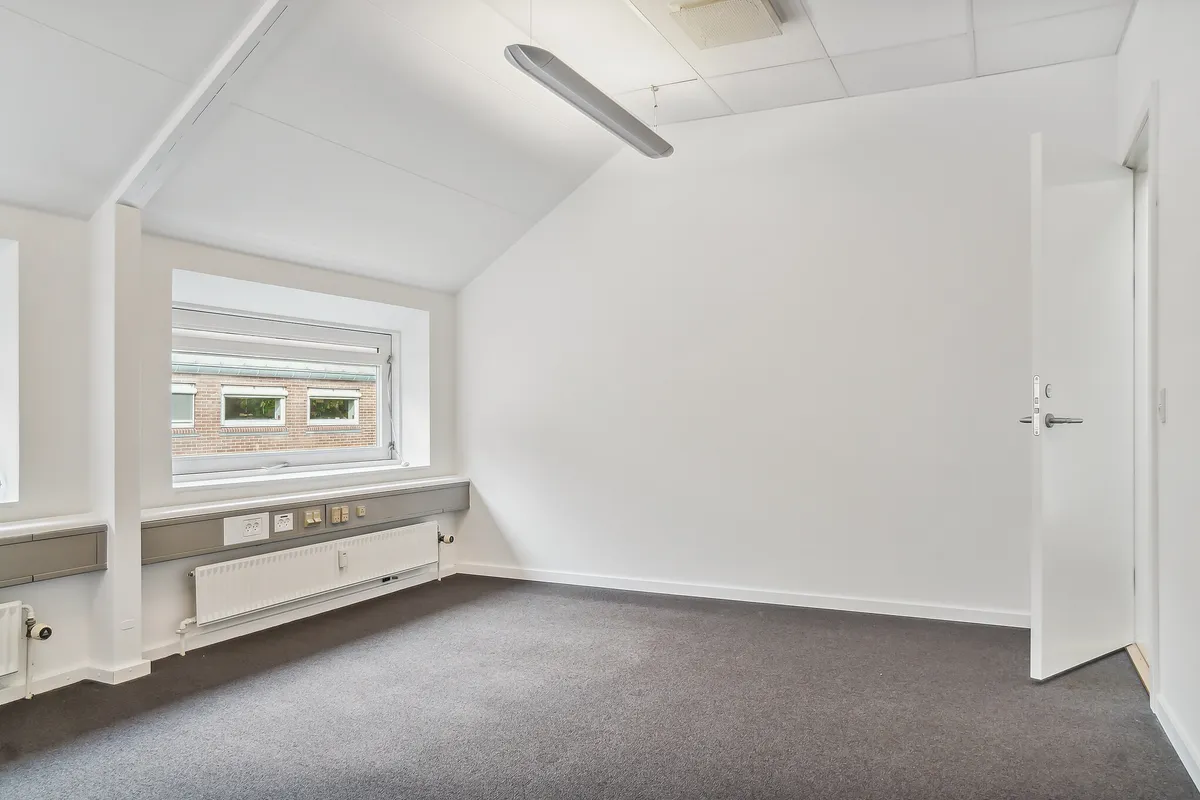 Lyserum med gulvtæppe og radiatorer langs væggen. Vindue med udsigt til bygning med røde mursten og to mindre ruder. Åben