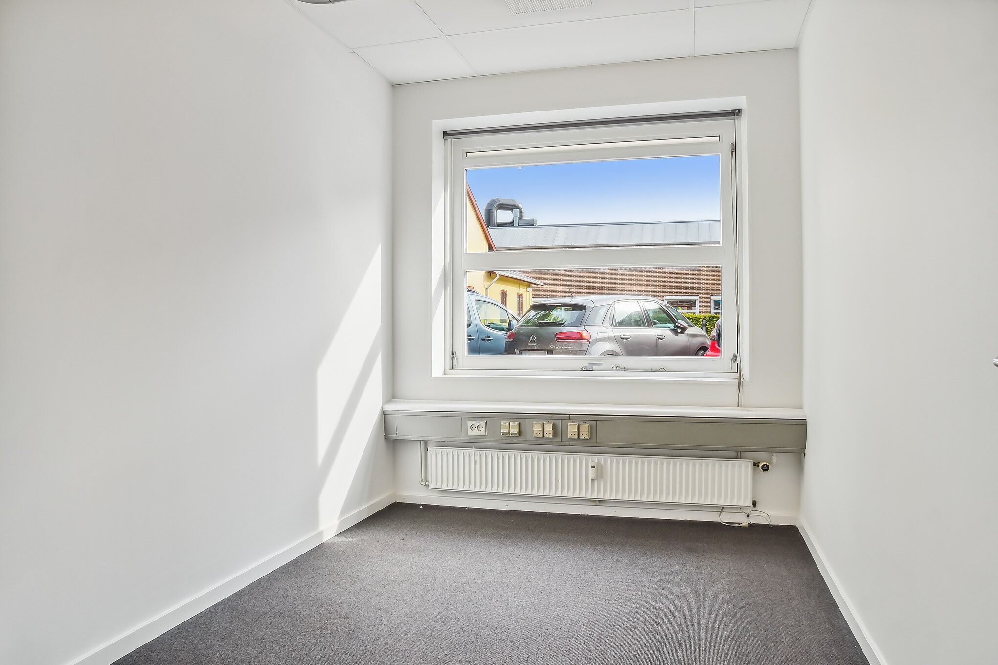 Ledig lokale med en stor vinduesfront, der vender mod en parkeringsplads med to biler. Væggene er hvide, og der er et