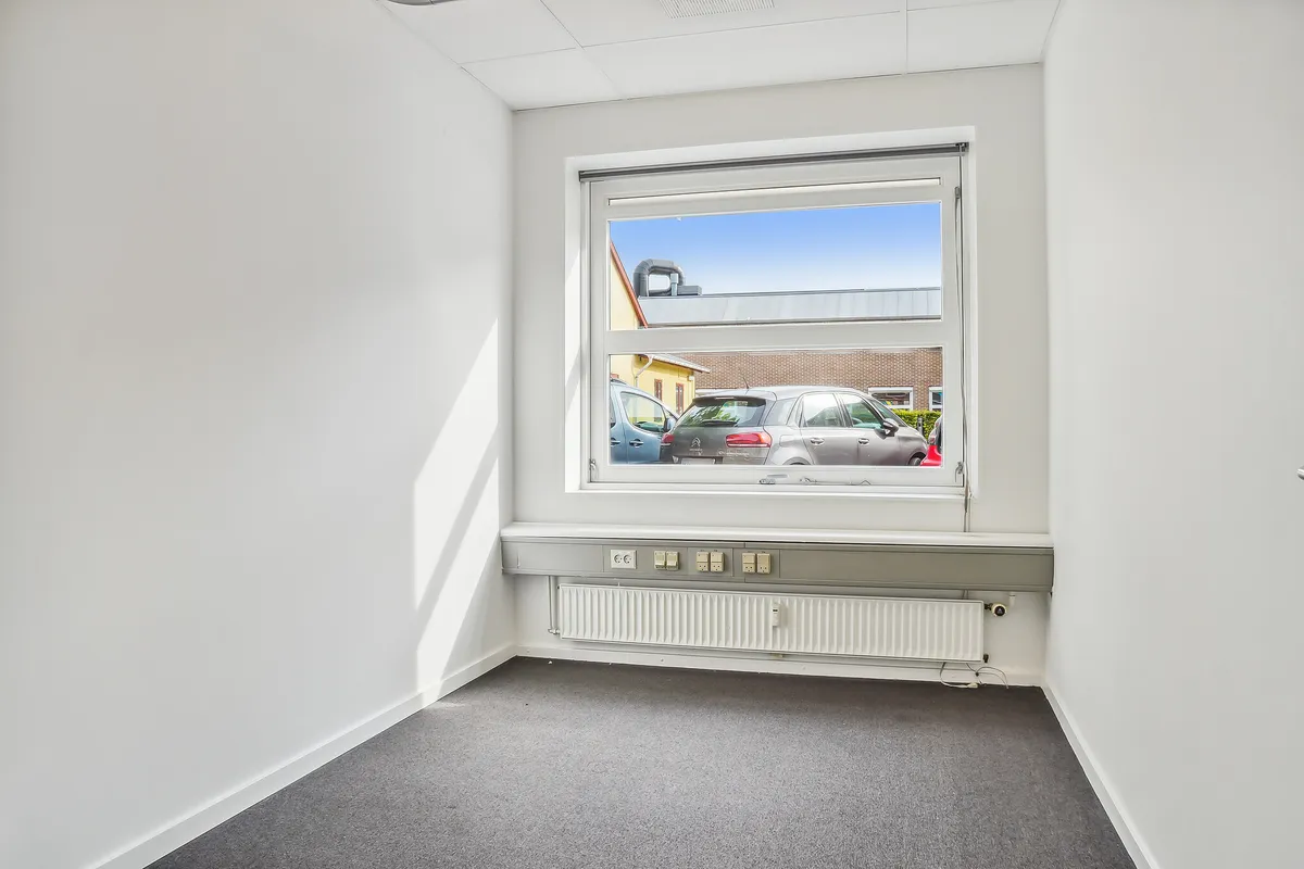 Ledig lokale med en stor vinduesfront, der vender mod en parkeringsplads med to biler. Væggene er hvide, og der er et
