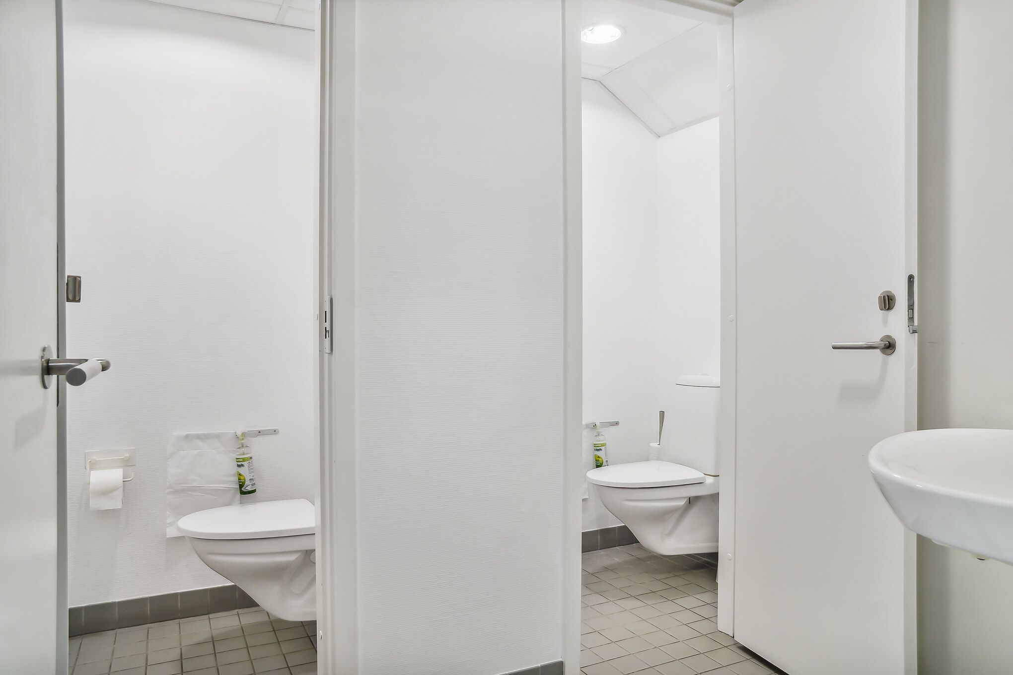 To toiletrum med hvide toiletter, håndvask, toiletpapir og desinfektionsmiddel samt et toilet med en lukket dør i et