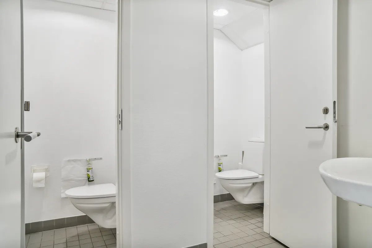 To toiletrum med hvide toiletter, håndvask, toiletpapir og desinfektionsmiddel samt et toilet med en lukket dør i et