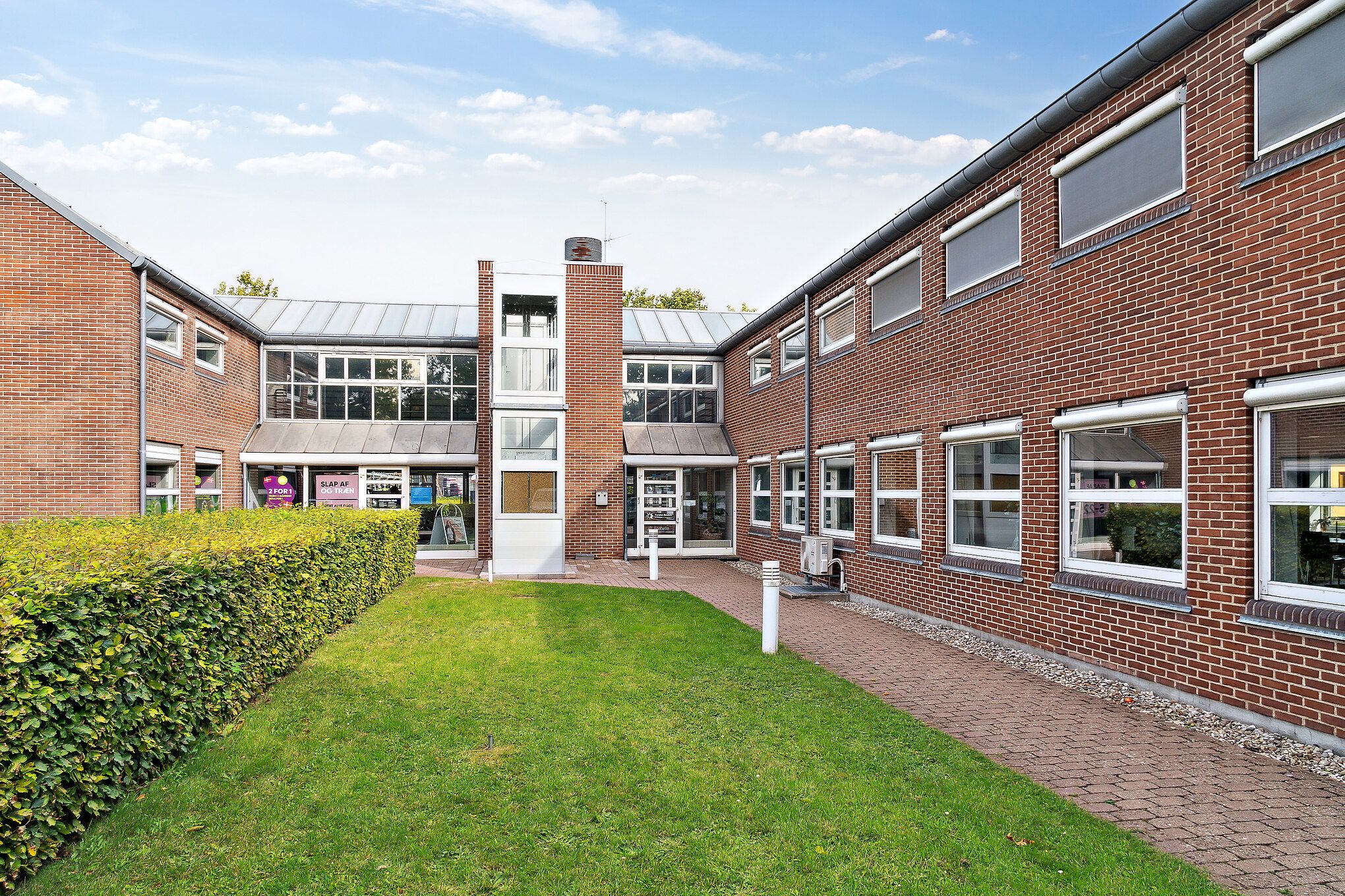 En række lokaler med rød mursten og store vinduer samt en grøn plæne og en buskehæk