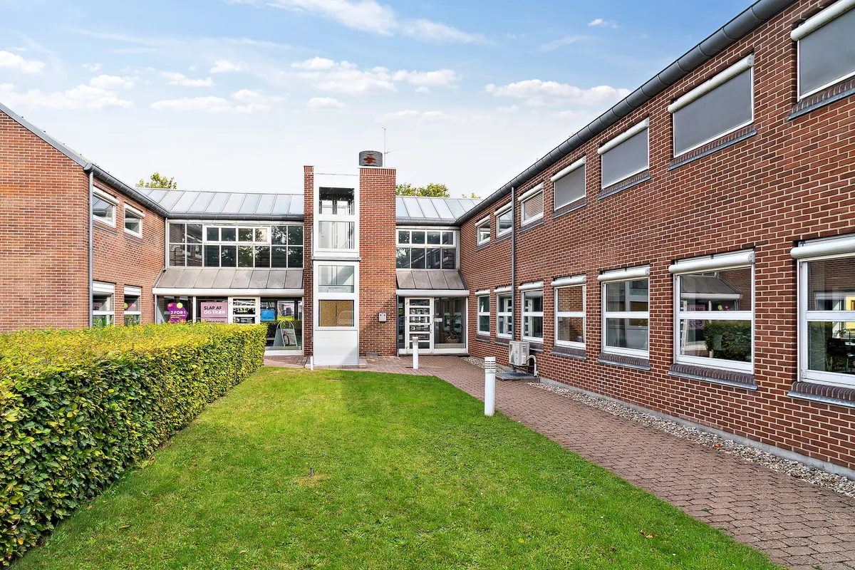 En række lokaler med rød mursten og store vinduer samt en grøn plæne og en buskehæk