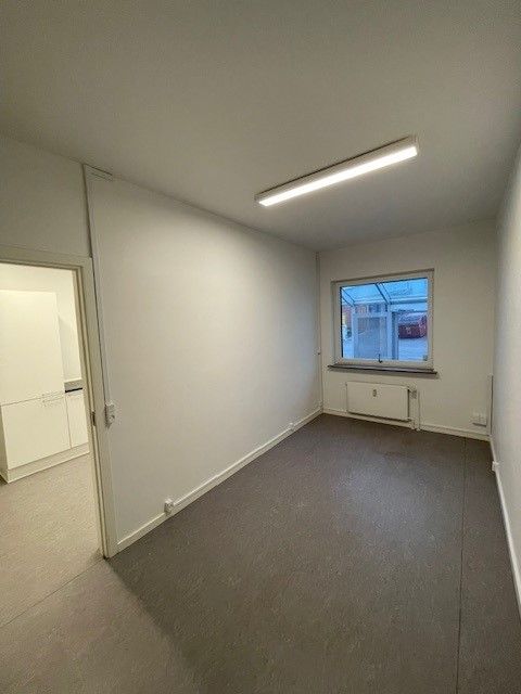 En tom rum med hvide vægge, lysegult gulv og en hvid dør der fører til et mindre rum med en hvid køleskab og et vindue