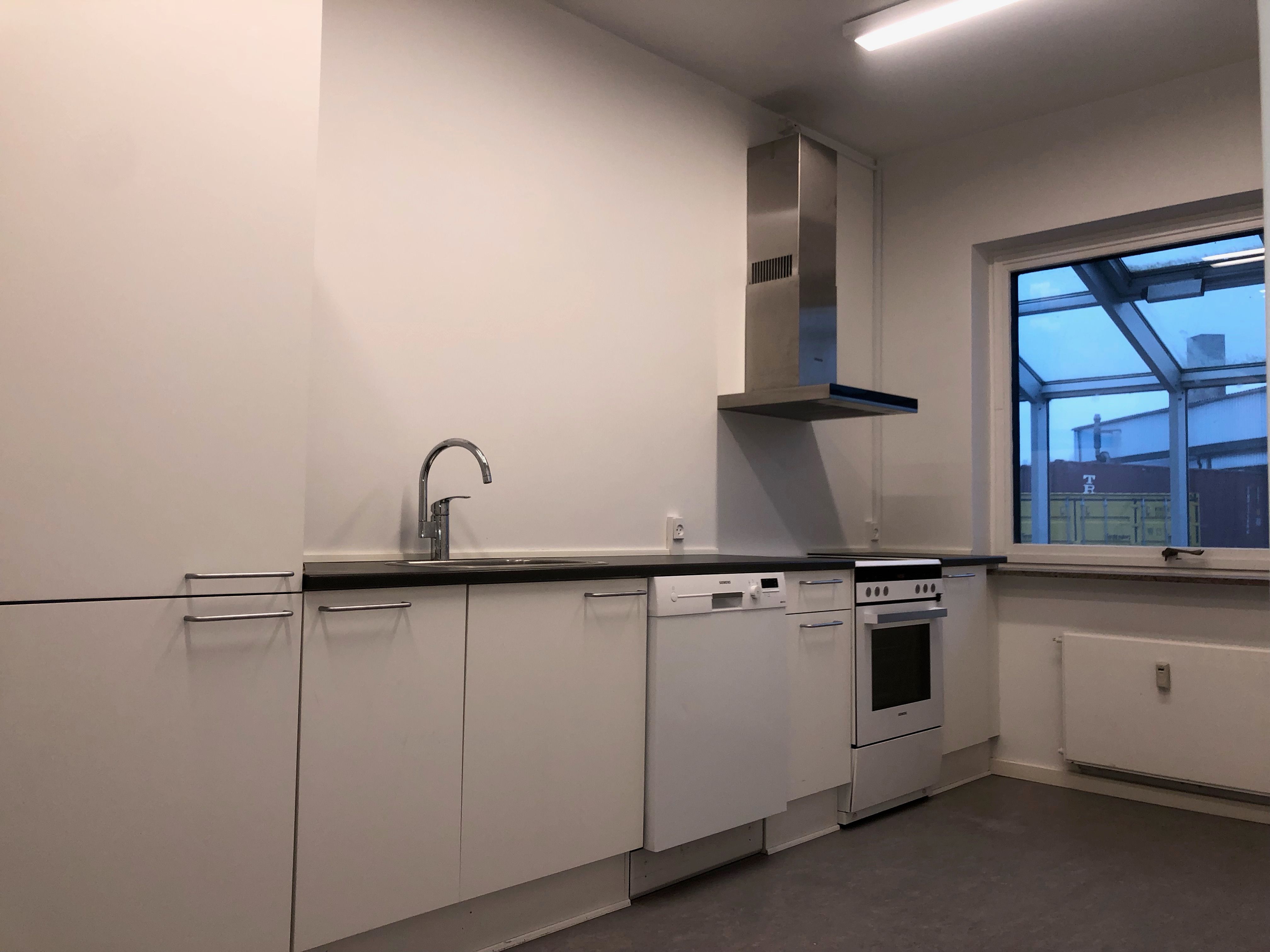 Et rent og moderne køkken med hvide vægge, lysegult lys, indbygget komfur, vaskemaskine, komfur og en høj vinduesglas og