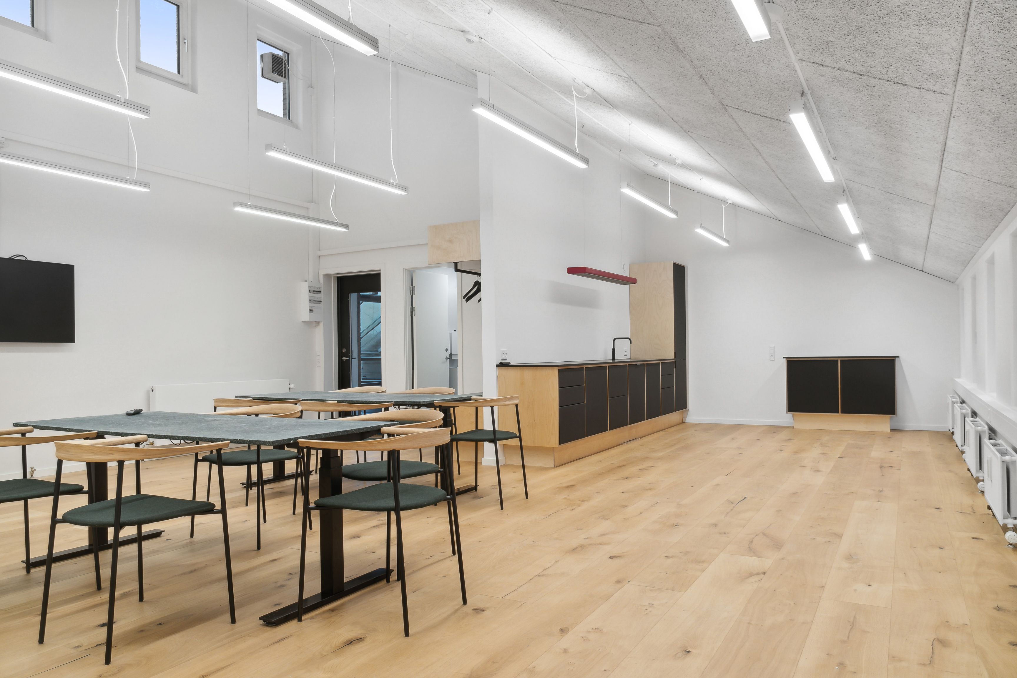 Sorte og hvide stole omkring grønne borde. Sorte højtalere og lysekroner. En køkkenvæg med en hængeklædeskab. Et køkken med