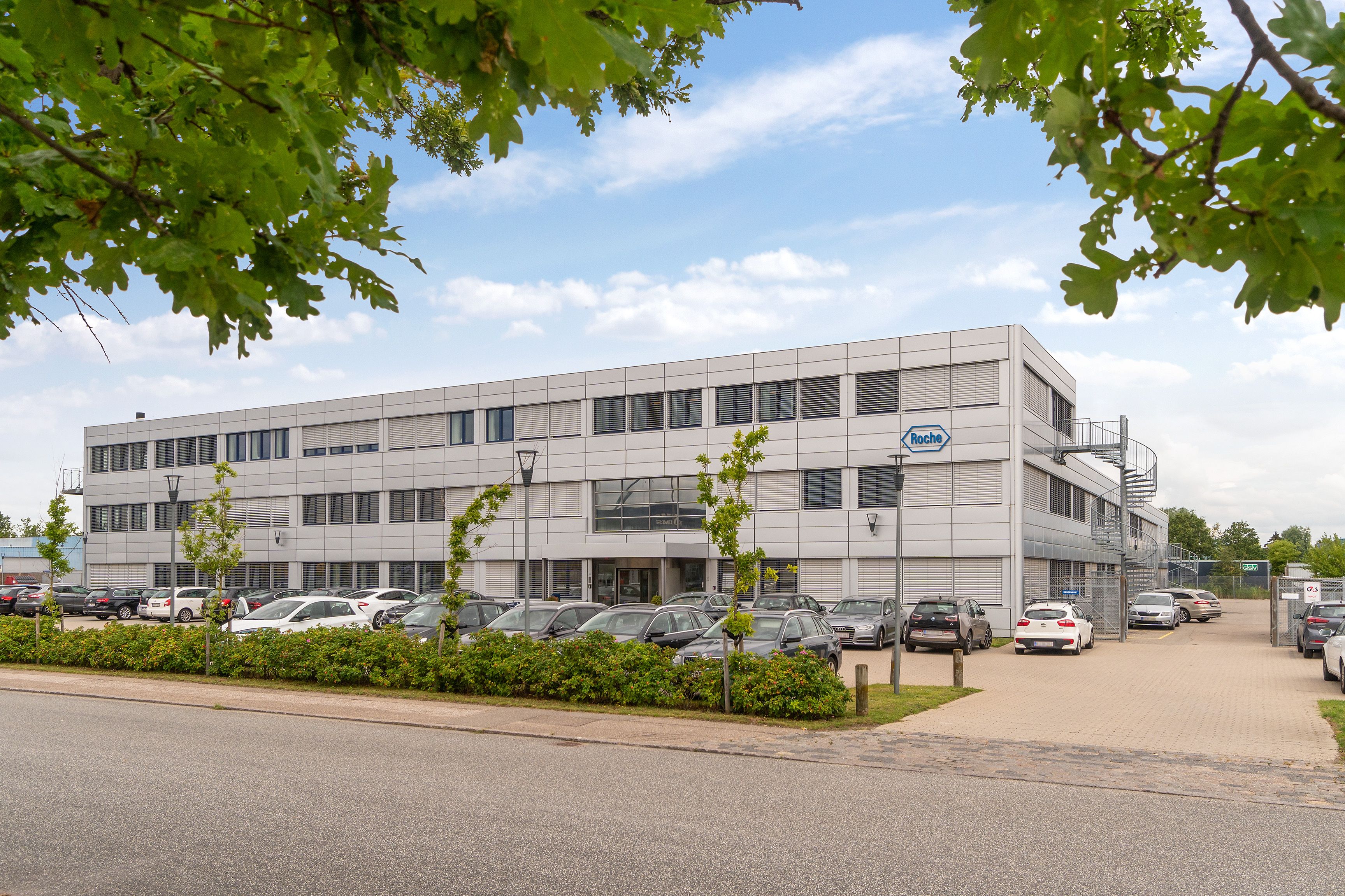 Store kontorer med parkeringsplads for biler og grønne planter og træer. Et rødt Roche logo er synligt på bygningen.