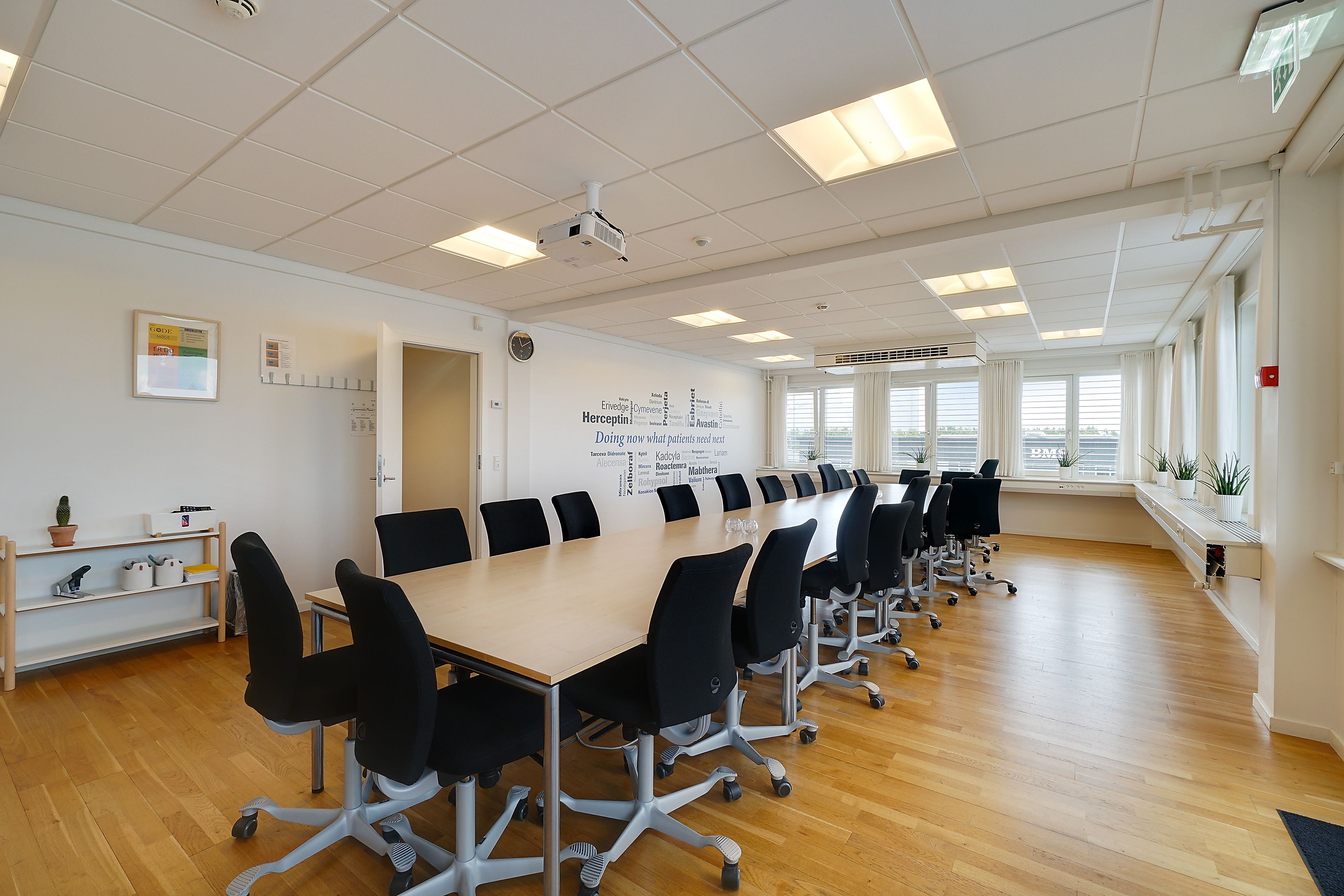 Mødeværelse med trægulv, projektor og en stor bord. Væggen er hvid med plakat og ure. Vinduerne er lukkede med persienner.