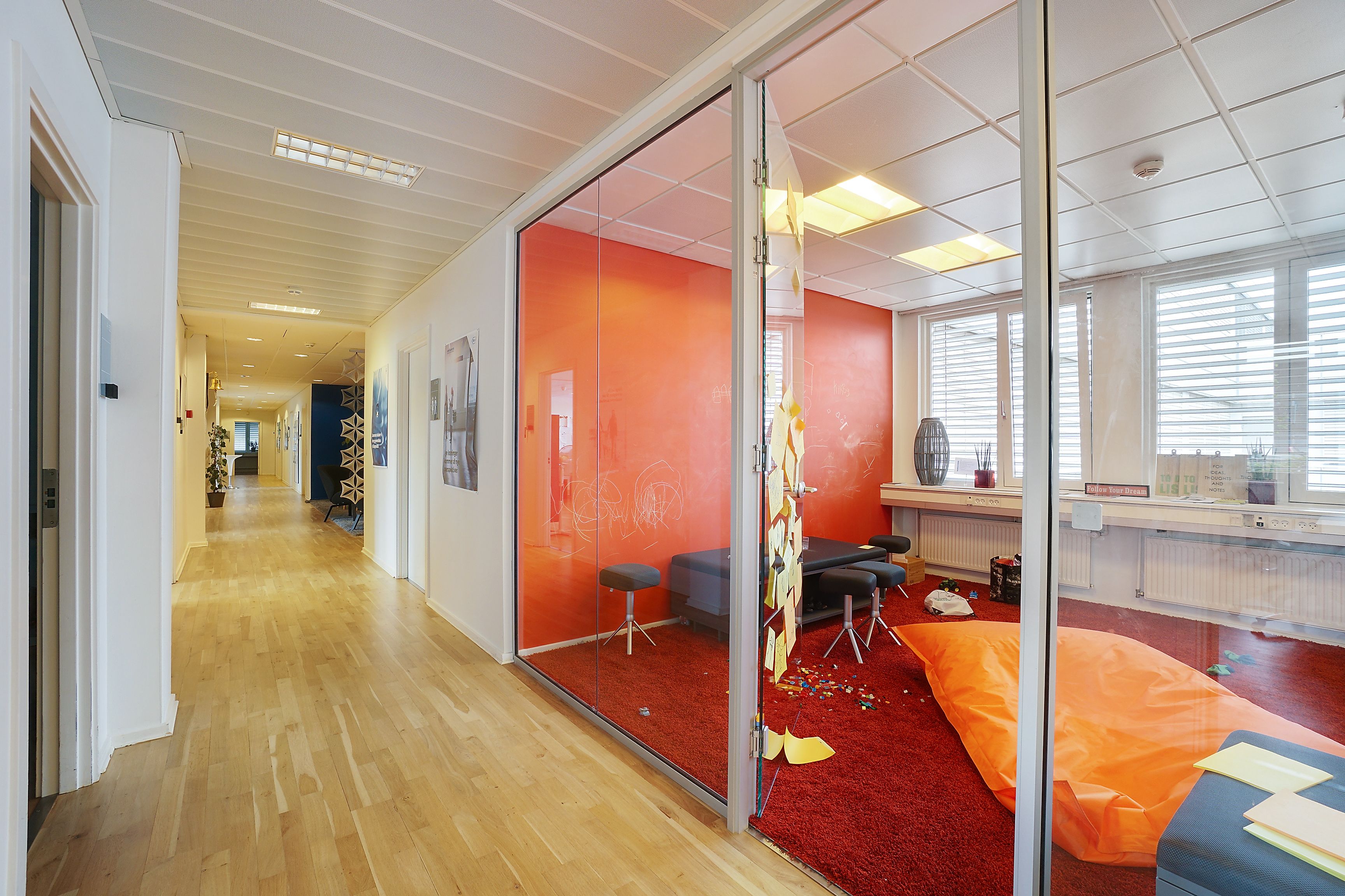 Flot indretning med lysegul trægulv, rødt gulvtæppe, glasvægge og orange puder samt stol med højt sæde.