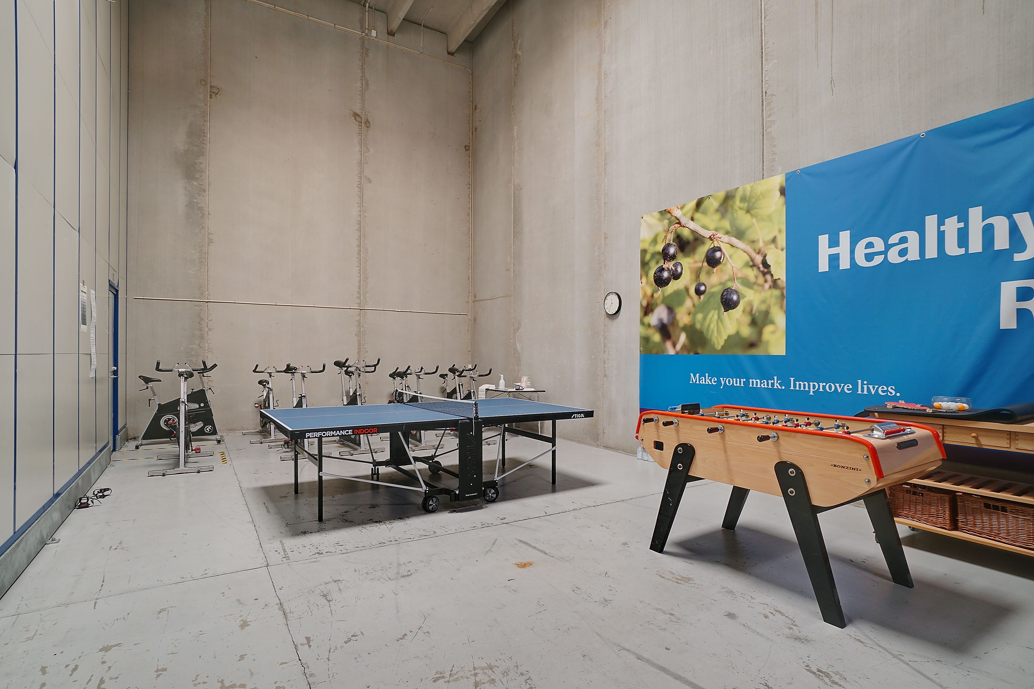 Spillelokale med bordtennisbord og fodboldbord med mange fitnessapparater i baggrunden samt en plakat med teksten Healthy