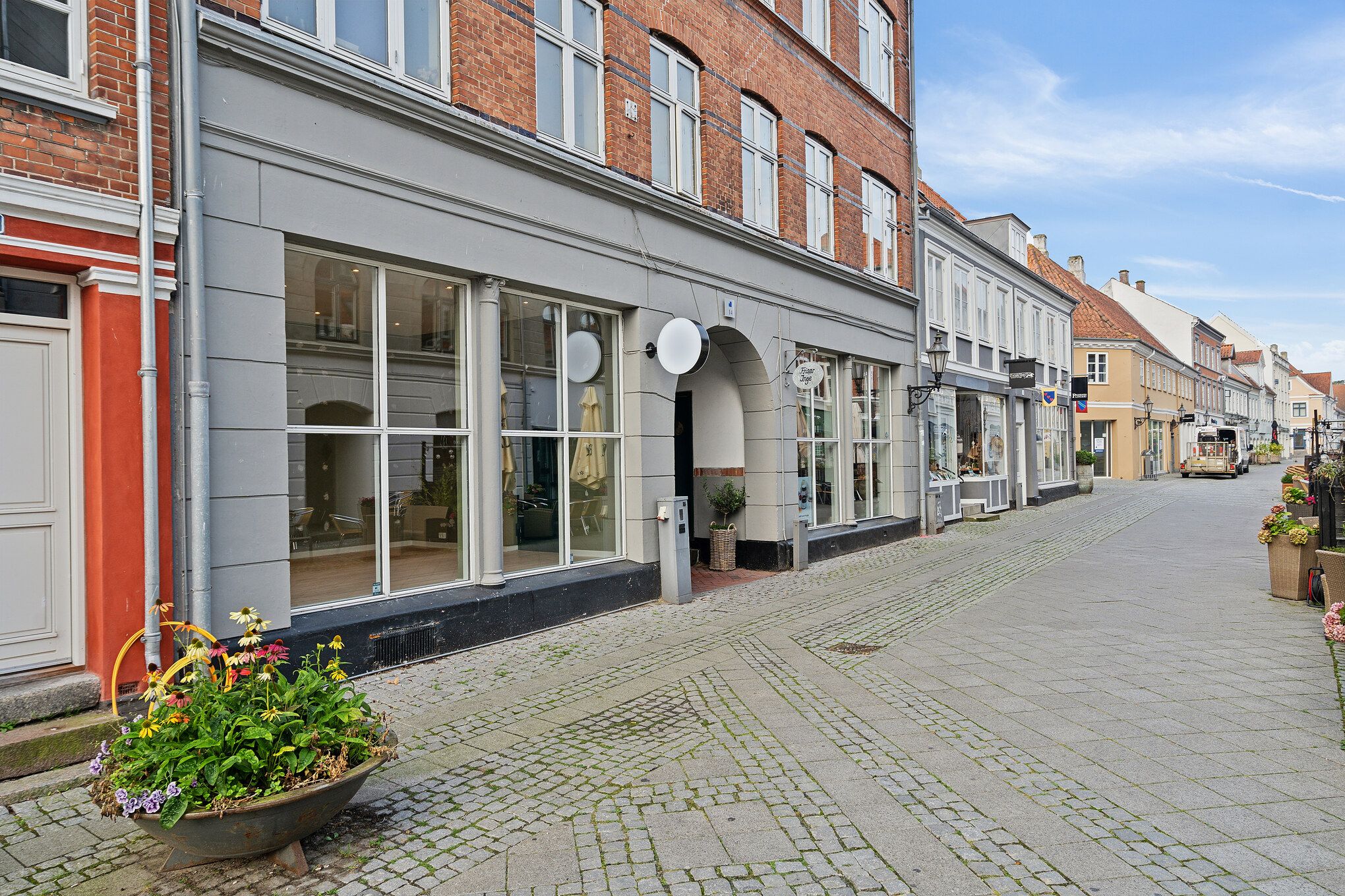 Gågade i Nyborg med butikker og blomsterarrangementer. Butikker med vinduer og døre. Bagved er en bus og bygninger.