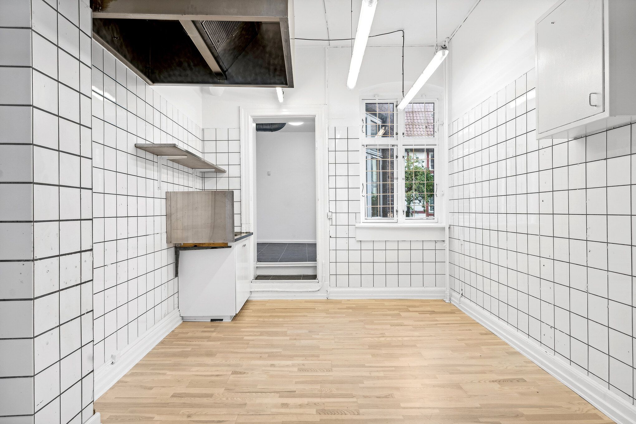 Hjørne af et køkken med hvide murfliser, to hængeskabe og lyse pærer på loftet. Et vinduesparti med dobbeltruder giver